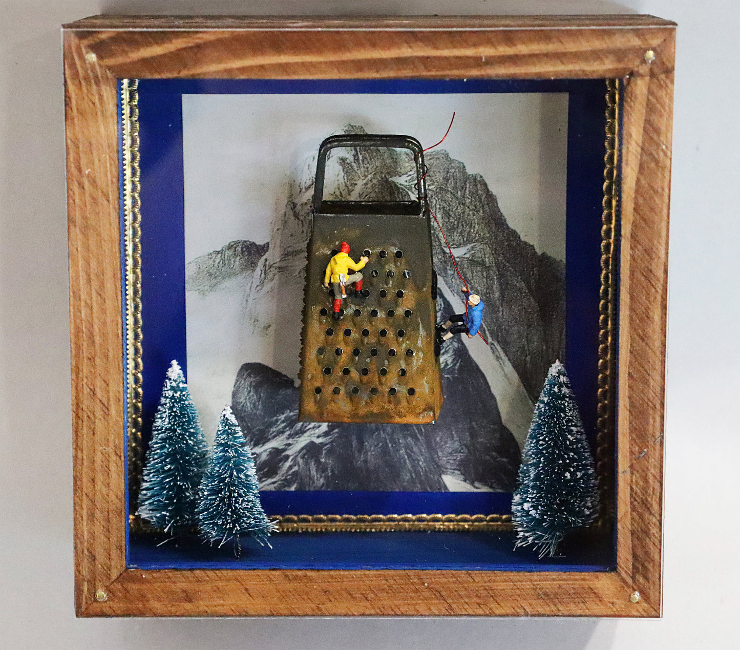 Original Objekt - Assemblage von Elisabeth Dishur, Titel „Der Bergsteiger“ – Objektkiste mit Miniatur-Figuren: Zwei Bergsteiger erklimmen eine Küchenreibe, darunter 3 verschneite Tannen, im Hintergrund eine Bergspitze. Eingefasst in eine Holzkiste, mit Plexiglas versehen - Objektcollage, erhältlich bei Galerie Waldbach - What The Artist Made