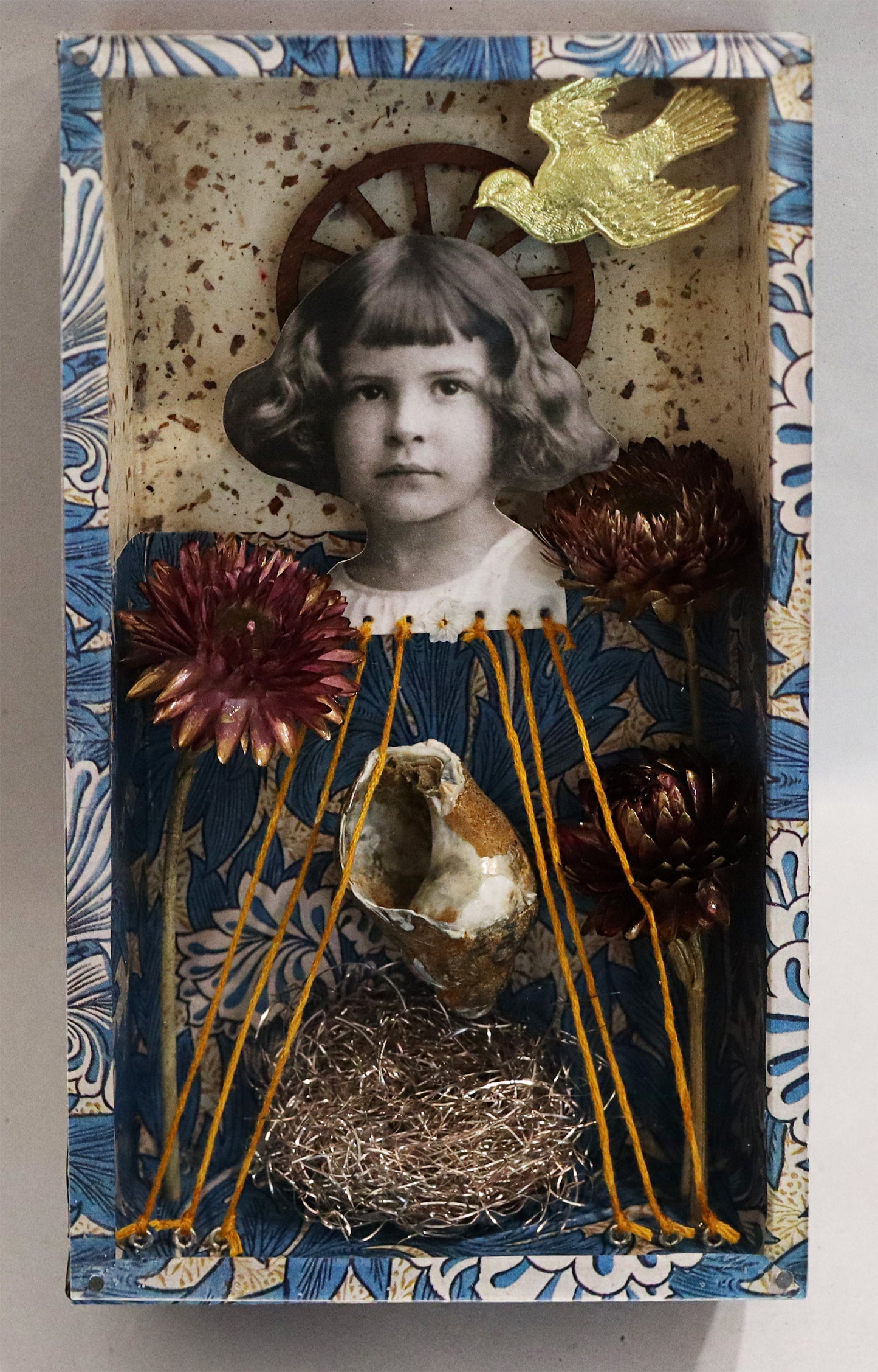 Original Objekt - Assemblage von Elisabeth Dishur, Titel „Minikiste“ – Objektkiste mit Gegenständen und Papiercollagen: Vintage-Porträt eines Mädchens, mit Fäden verbunden mit Boden der Kiste, darüber eine goldene Taube fliegend. Umfasst von Trockenblumen, Schneckenhaus und Vogelnest, Floraler Hintergrund in Blautönen. Eingefasst in eine Holzkiste, mit Plexiglas versehen - Objektcollage, erhältlich bei Galerie Waldbach - What The Artist Made