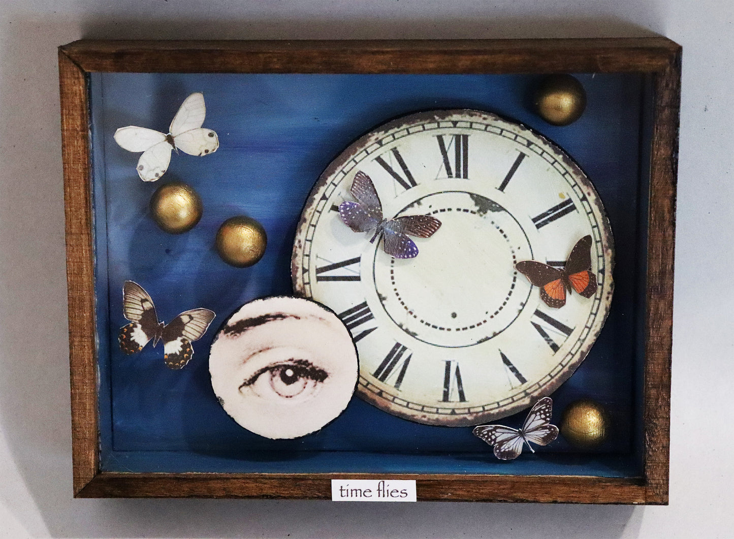 Original Objekt - Assemblage von Elisabeth Dishur, Titel „time flies“ – Objektkiste mit Papiercollagen und Gegenständen: Ein Ziffernblatt aus Papier, das die Kiste dominiert, daneben aus Papier ein rundes Auge. Fünf Schmetterlinge aus Papier und 4 goldene Kugeln sind darum angeordnet, der Hintergrund ist blau und der Titel steht unten am Rand auf dem Holz. Eingefasst in eine Holzkiste, mit Plexiglas versehen - Objektcollage, erhältlich bei Galerie Waldbach - What The Artist Made