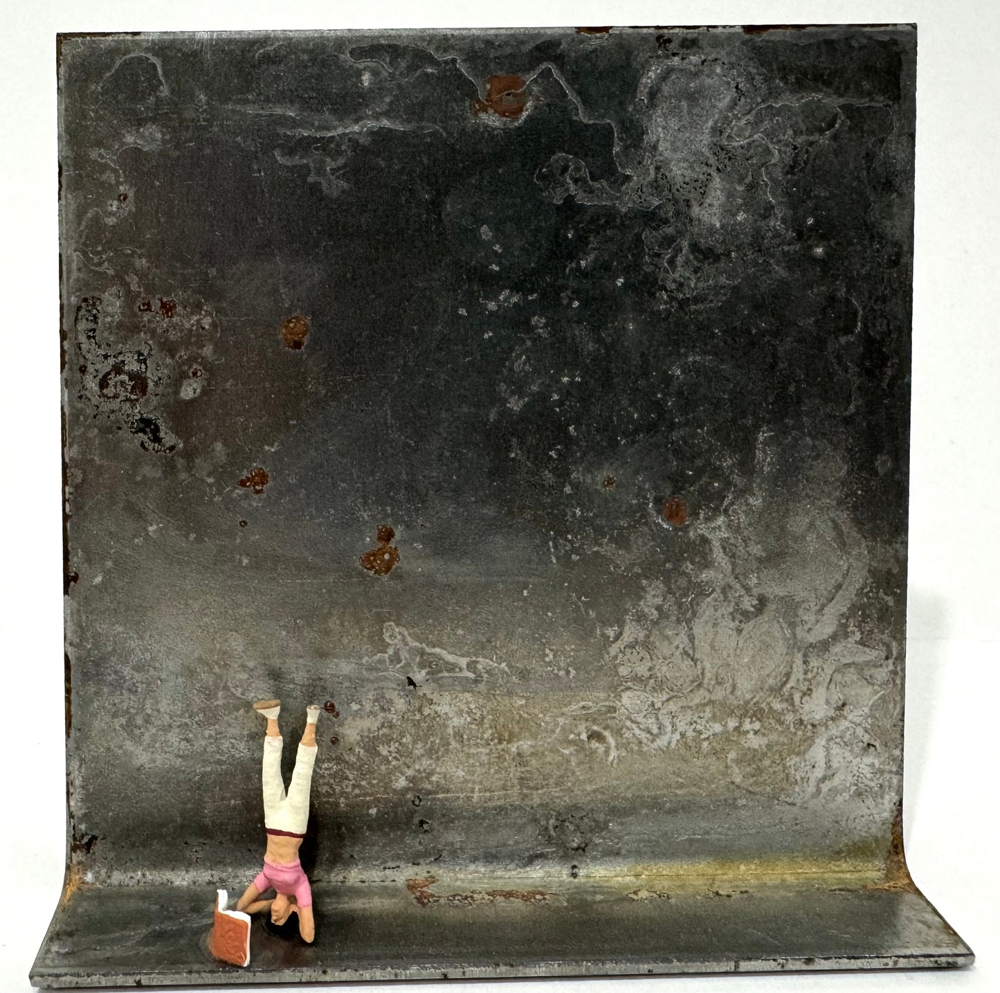 Original Objekt - Assemblage von Katrin Schramm, Titel „ 180°“ – Metallwinkel mit Miniatur-Figur: Frau in Kopfstand links auf dem Winkel stehend, vor ihr ein Buch aufgeklappt stehend. Dahinter das Metall des Winkels als Hintergrund. - Objektcollage, erhältlich bei What The Artist Made