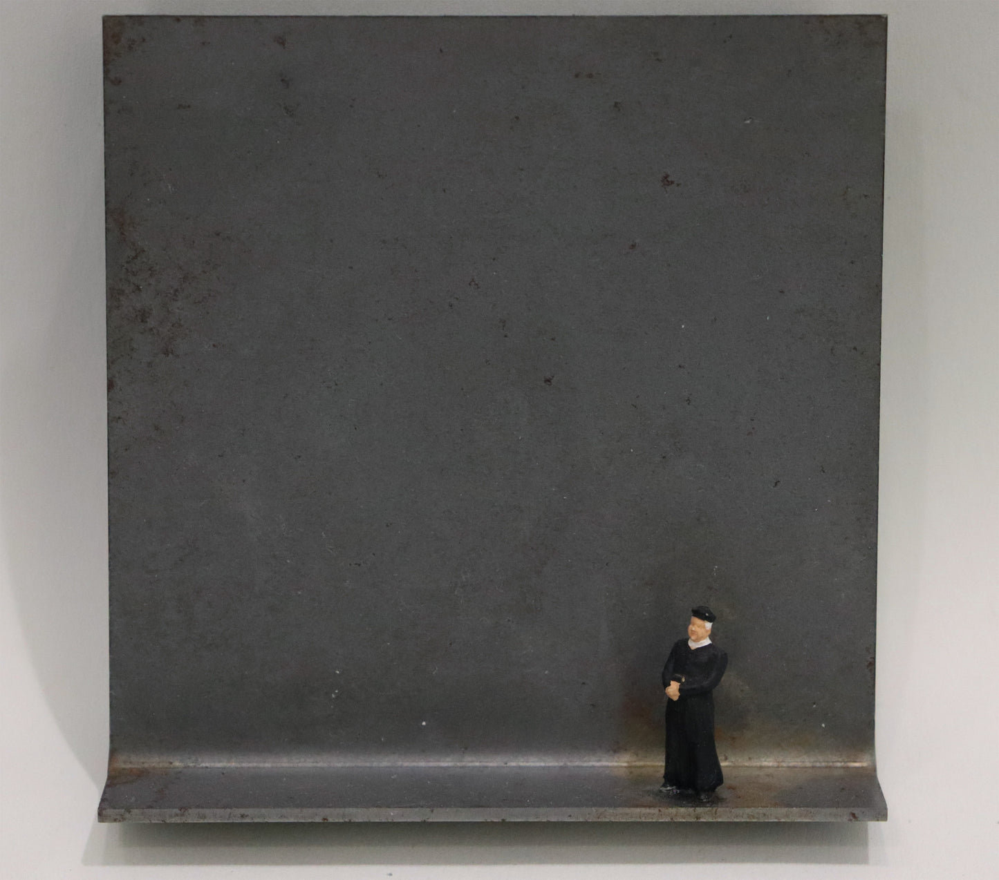 Original Objekt - Assemblage von Katrin Schramm, Titel „Der Priester, immer der Priester.“ – Metallwinkel mit Miniatur-Figur: Priester in schwarzem Habit mit Hut stehend, Hände gefaltet. Dahinter das Metall des Winkels mit Struktur, als Hintergrund. - Objektcollage, erhältlich bei Galerie Waldbach - What The Artist Made
