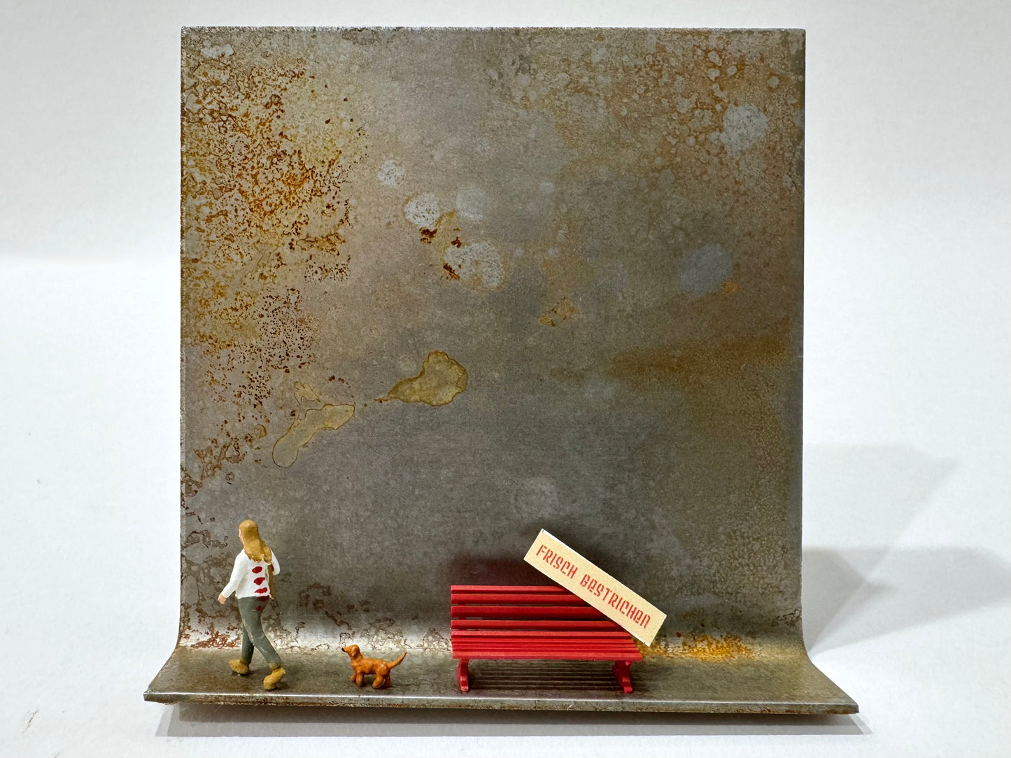Original Objekt - Assemblage von Katrin Schramm, Titel „Gelackt“ – Metallwinkel mit Miniatur-Figuren: eine Frau, aus der Szene gehend am linken Rand des Metallwinkels, hinter ihr ein kleiner brauner Hund, daneben eine rote Bank mit. „frisch gestrichen“-Schild. Dahinter das Metall des Winkels als Hintergrund. - Objektcollage, erhältlich bei What The Artist Made