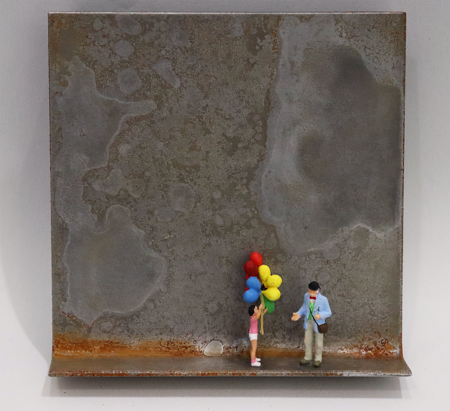 Original Objekt - Assemblage von Katrin Schramm, Titel „Lass sie fliegen...“ – Metallwinkel mit Miniatur-Figuren: Ein Mädchen mit Luftballons an einem Seil stehend, neben ihr ein älterer Herr mit Hut und Fliege. Dahinter das Metall des Winkels mit Struktur, als Hintergrund. - Objektcollage, erhältlich bei Galerie Waldbach - What The Artist Made