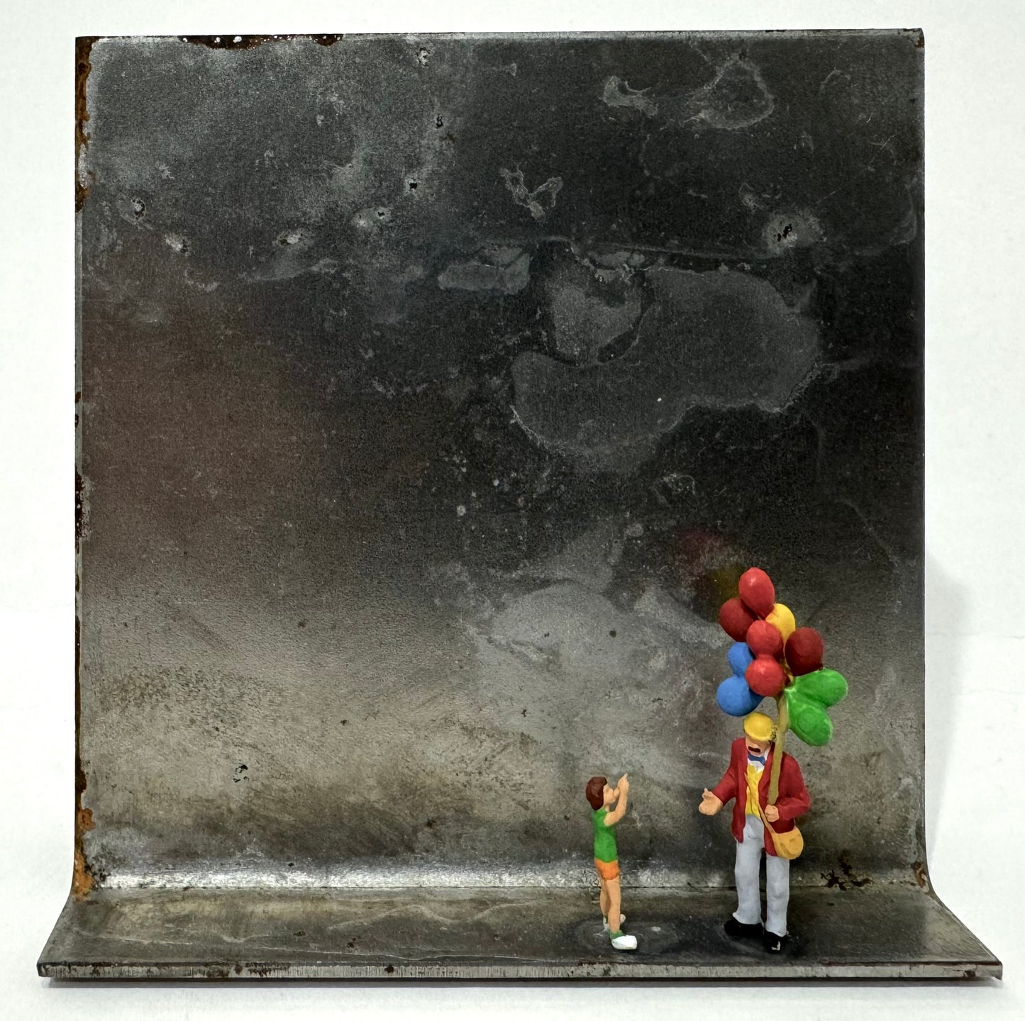 Original Objekt - Assemblage von Katrin Schramm, Titel „Lass sie fliegen…“ – Metallwinkel mit Miniatur-Figuren: Ballonverkäufer auf dem Winkel stehend, daneben ein Kind, welches auf die Luftballons zeigt, die er in der Hand festhält. Dahinter das Metall des Winkels als Hintergrund. - Objektcollage, erhältlich bei What The Artist Made