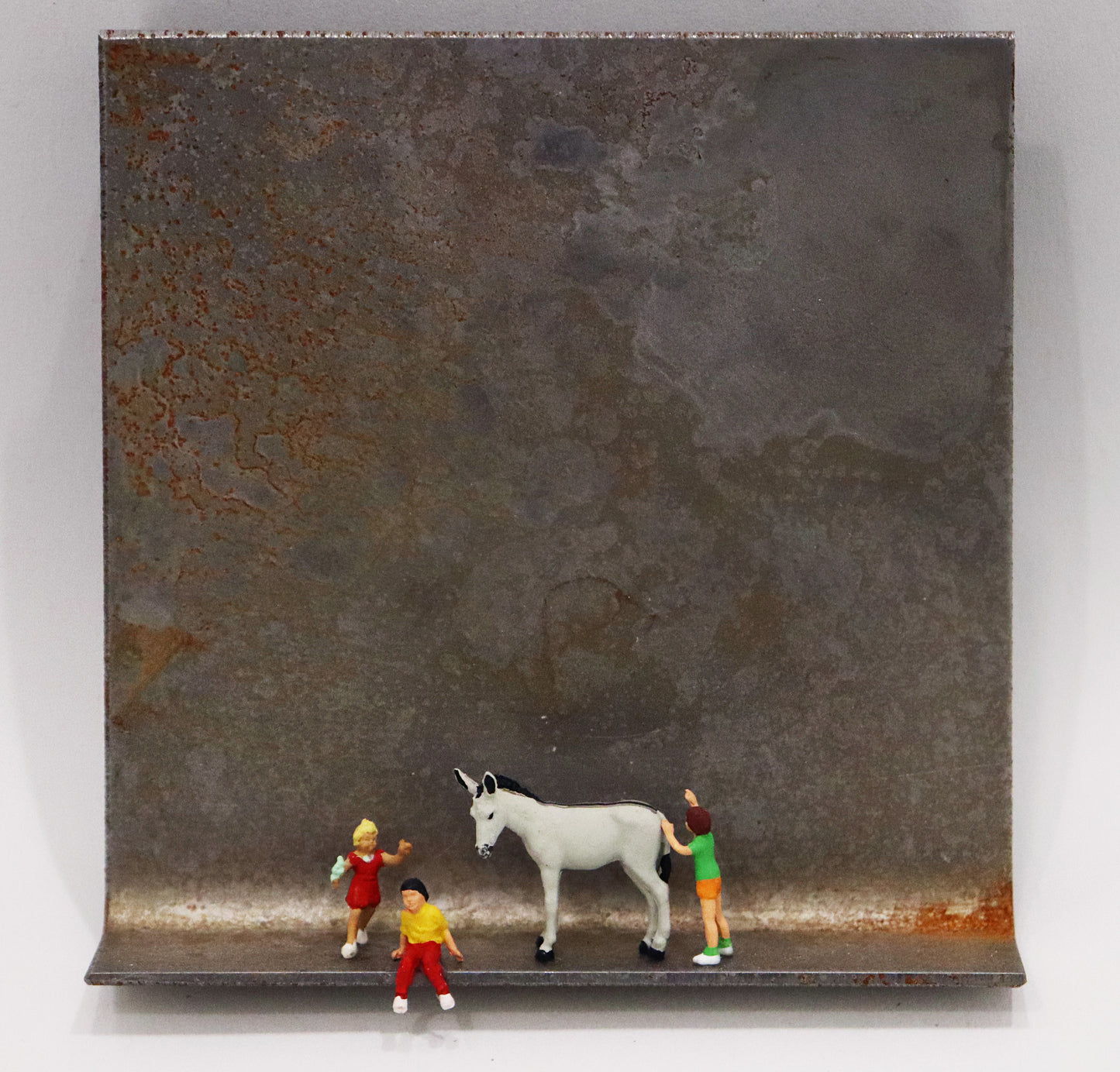 Original Objekt - Assemblage von Katrin Schramm, Titel „Mit Eselsgeduld...“ – Metallwinkel mit Miniatur-Figuren: Mittig stehend/sitzend zwei Kinder, daneben ein weißer Esel, rechts hinter diesem ein weiteres Kind stehend, vermeintlich den Esel schiebend. Dahinter das Metall des Winkels mit Struktur, als Hintergrund. - Objektcollage, erhältlich bei Galerie Waldbach - What The Artist Made