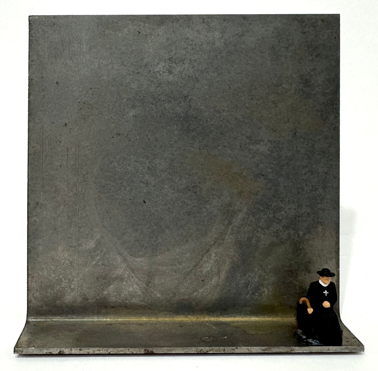 Original Objekt - Assemblage von Katrin Schramm, Titel „Sonderzug“ – Metallwinkel mit Miniatur-Figur: ein Priester/Pfarrer, am rechten Rand des Metallwinkels sitzend. Dahinter das Metall des Winkels als Hintergrund. - Objektcollage, erhältlich bei What The Artist Made