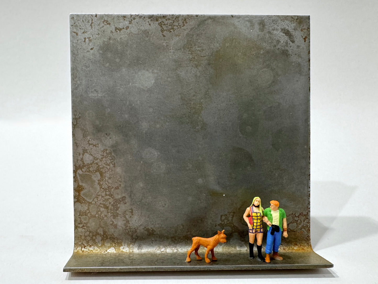 Original Objekt - Assemblage von Katrin Schramm, Titel „Wir sind jetzt zu dritt“ – Metallwinkel mit Miniatur-Figuren: ein Paar auf dem Winkel stehend, neben beiden ein brauner Hund, dahinter das Metall des Winkels als Hintergrund. - Objektcollage, erhältlich bei What The Artist Made