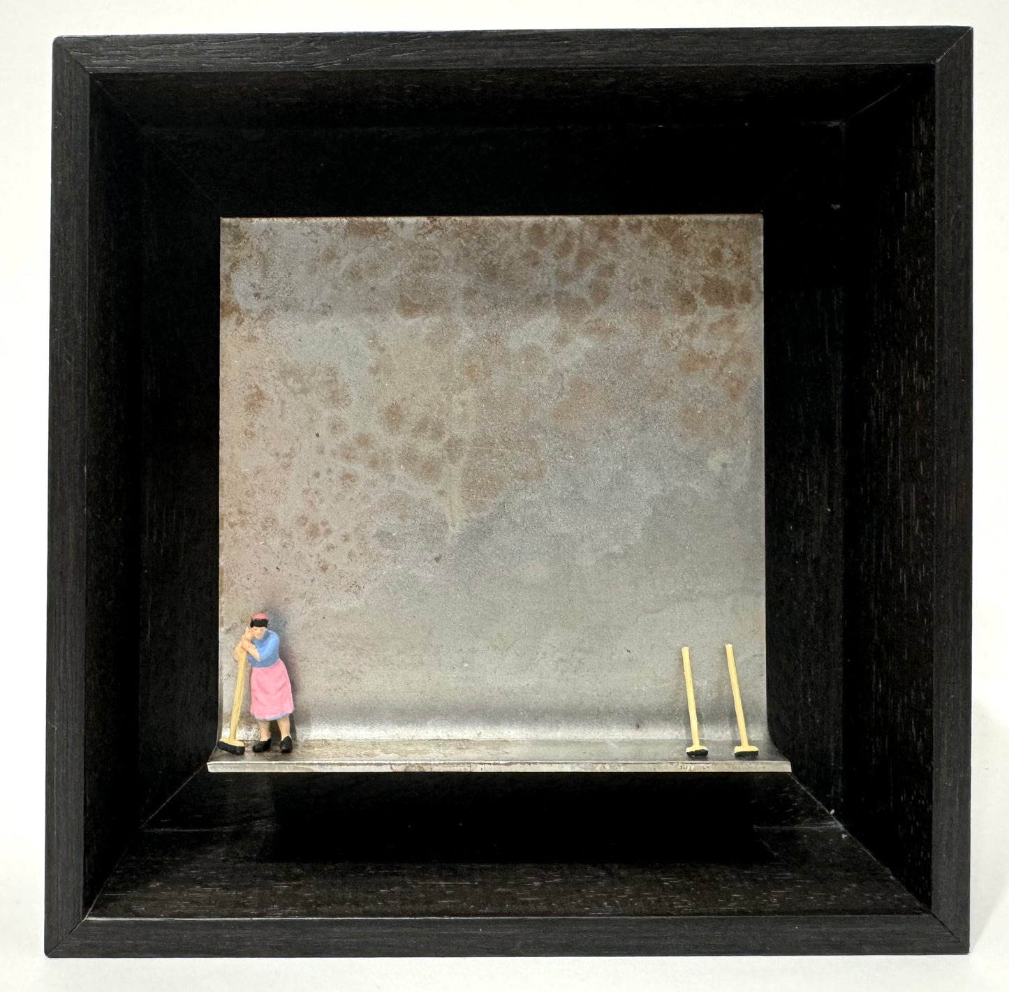 Original Objekt - Assemblage von Katrin Schramm, Titel „das bißchen Haushalt…“ – Metallwinkel mit Miniatur-Figur: Frau links am Rand des Winkels stehend, stützt sich auf Besen. Rechts am Rand zwei weitere Besen gegen den Hintergrund des Metallwinkels gelehnt. - Objektcollage, erhältlich bei What The Artist Made