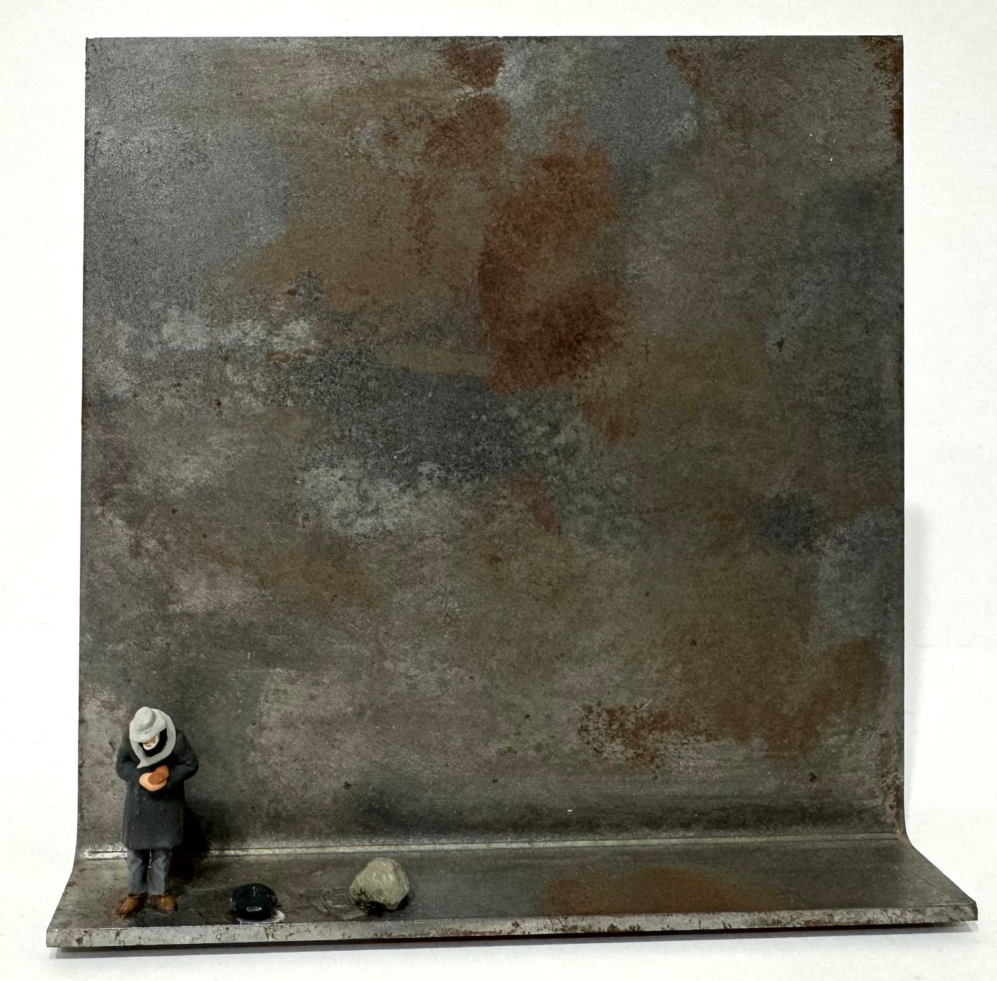 Original Objekt - Assemblage von Katrin Schramm, Titel „geben…“ – Metallwinkel mit Miniatur-Figur: Person am linken Rand des Metallwinkels stehend, Kopf gebeugt, etwas in der Hand haltend. Neben dem Mann ein Gefäß liegend, evtl. ein Hut, daneben ein größerer Stein/Fels. Dahinter das Metall des Winkels als Hintergrund. - Objektcollage, erhältlich bei What The Artist Made