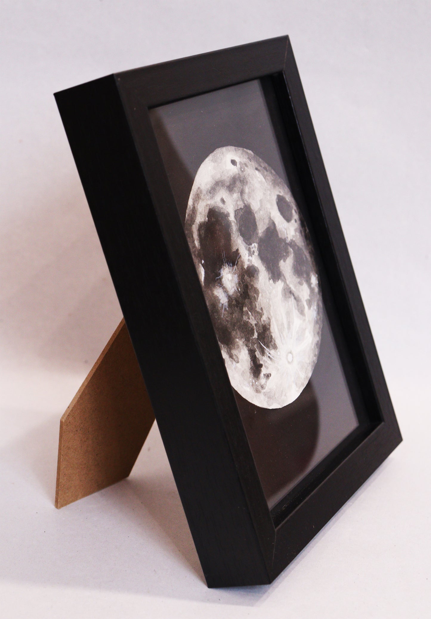 Original Aquarell-Malerei, Gemälde von Lisa Steinbrück, Titel „Luna“ – Darstellung des Mondes der Erde in Aquarell-Stil, schwarz-weiß und Grautöne auf schwarzem Grund, Aquarell auf Papier, gerahmt, schwarzer Rahmen, Seitenansicht auf den stehenden Rahmen - erhältlich bei Galerie Waldbach - What The Artist Made