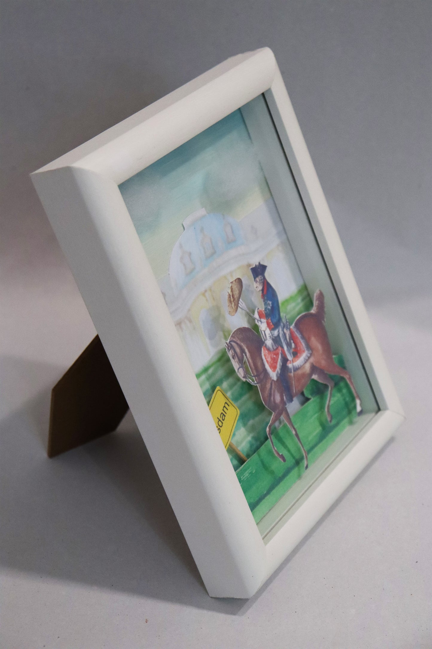 Collage nach einer Malerei von Nadine Conrad, Titel „Kartoffelfritze“ – Friedrich II auf Pferd reitend, er hält eine große Gabel wie eine Lanze, darauf steckt eine große Kartoffel. Links von ihm ist das Ortsschild von Potsdam, im Hintergrund das Schloss Sanssouci, Papiercollage, gerahmt - Seitenansicht auf den stehenden weißen Rahmen. Erhältlich bei Galerie Waldbach - What The Artist Made