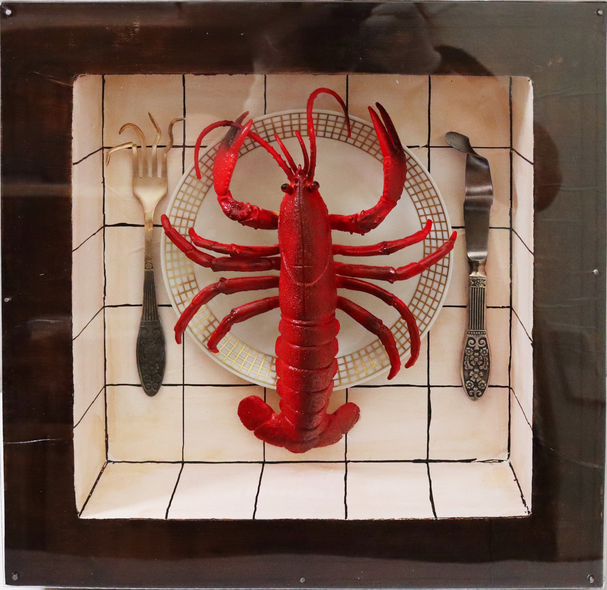 Original Objekt - Assemblage von Elisabeth Dishur, Titel „Hummer“ – Objektkiste mit Gegenständen: Ein roter Hummer aus Kunststoff liegt auf einem weißen Teller mit Goldrand, links und rechts liegen Gabel und Messer, die Zinken und Schneide verbogen. Der Hintergrund ist weiß mit schwarzen Quadraten -  an Fließen erinnernd. Eingefasst in eine Holzkiste, mit Plexiglas versehen - Objektcollage, erhältlich bei Galerie Waldbach - What The Artist Made