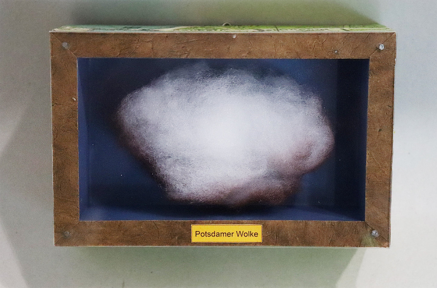 Original Objekt - Assemblage von Elisabeth Dishur, Titel „Potsdamer Wolke" – Objektkiste mit Wolke aus Watte, gefärbt. Blauer Hintergrund, Titel unten mittig auf dem Kistenrand. Eingefasst in eine Holzkiste, umseitig Papiercollage mit Stadtkarte, mit Plexiglas versehen - Objektcollage, erhältlich bei Galerie Waldbach - What The Artist Made