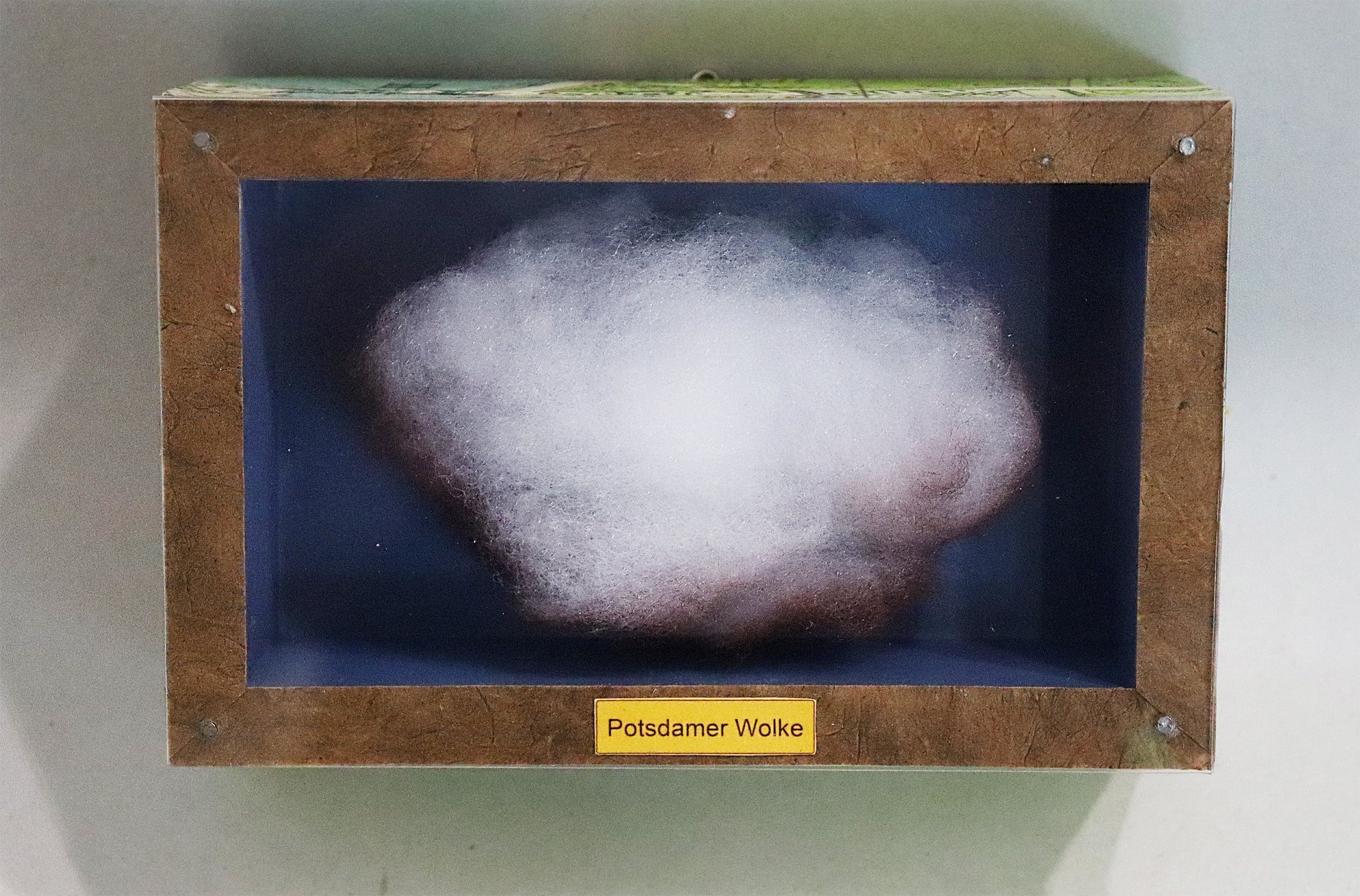 Original Objekt - Assemblage von Elisabeth Dishur, Titel „Potsdamer Wolke" – Objektkiste mit Wolke aus Watte, gefärbt. Blauer Hintergrund, Titel unten mittig auf dem Kistenrand. Eingefasst in eine Holzkiste, umseitig Papiercollage mit Stadtkarte, mit Plexiglas versehen - Objektcollage, erhältlich bei Galerie Waldbach - What The Artist Made