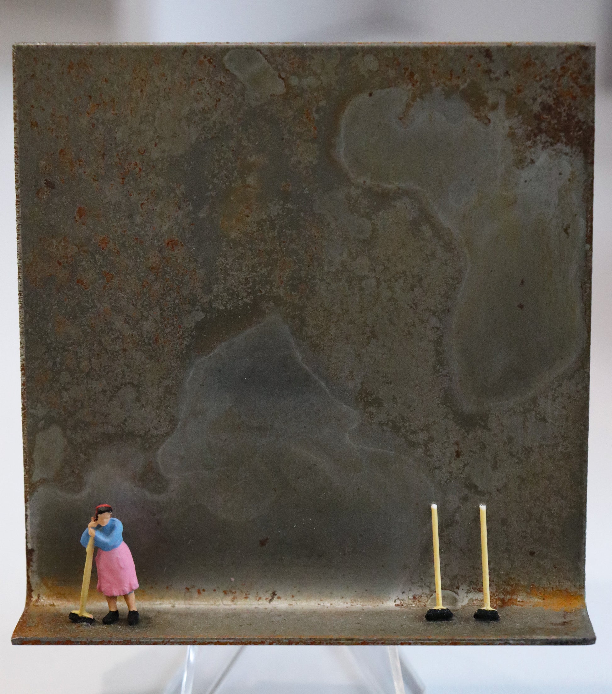 Original Objekt - Assemblage von Katrin Schramm, Titel „Das bisschen Haushalt...“ – Metallwinkel mit Miniatur-Figur: Eine Frau steht in Hausarbeitskleidung am linken Rand des Winkels und stützt sich an einen Besen. Am rechten Rand stehen zwei Kehrbesen angelehnt. Dahinter das Metall des Winkels mit Struktur, als Hintergrund. Objektcollage, erhältlich bei Galerie Waldbach - What The Artist Made