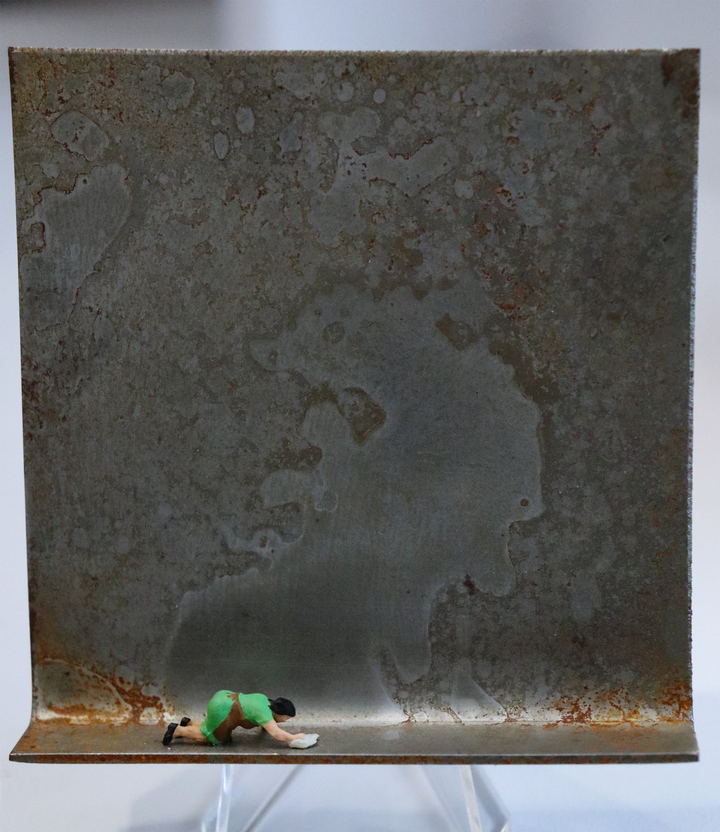 Original Objekt - Assemblage von Katrin Schramm, Titel „Ich möchte ein Glanz sein...“ – Metallwinkel mit Miniatur-Figur: Eine Frau kniet in Haushaltskleidung am Boden des  Winkels und wischt diesen mit einem Lappen. Dahinter das Metall des Winkels mit Struktur, als Hintergrund. Objektcollage, erhältlich bei Galerie Waldbach - What The Artist Made