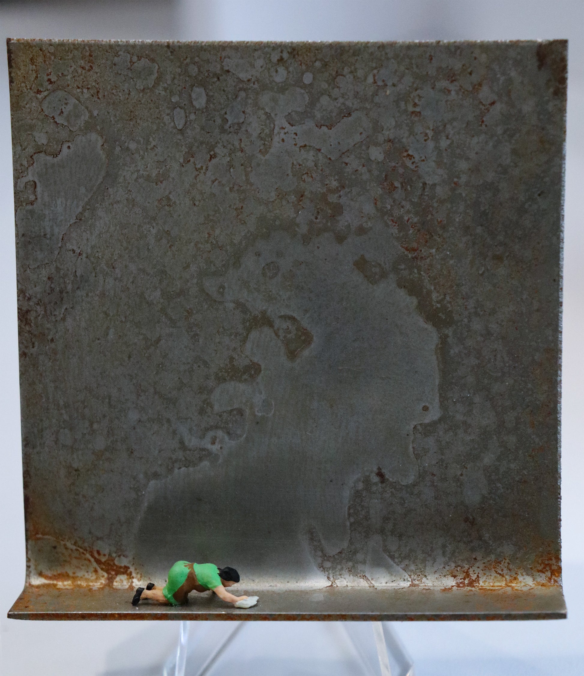 Original Objekt - Assemblage von Katrin Schramm, Titel „Ich möchte ein Glanz sein...“ – Metallwinkel mit Miniatur-Figur: Eine Frau kniet in Haushaltskleidung am Boden des  Winkels und wischt diesen mit einem Lappen. Dahinter das Metall des Winkels mit Struktur, als Hintergrund. Objektcollage, erhältlich bei Galerie Waldbach - What The Artist Made