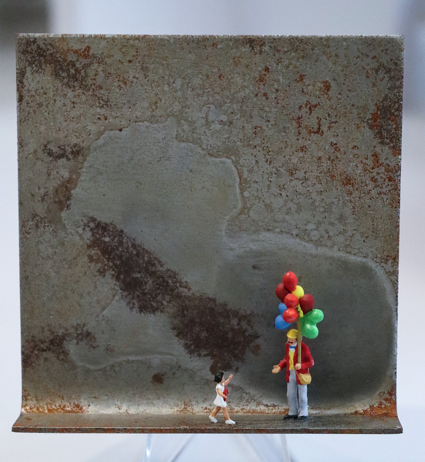 Original Objekt - Assemblage von Katrin Schramm, Titel „Lass sie fliegen...“ – Metallwinkel mit Miniatur-Figuren: Ein Luftballonverkäufer mit vielen bunten Ballons, stehend, hält die  Hand auf und schaut zu einem kleinen Mädchen im weißen Kleid, dass auf  ihn zuläuft. Dahinter das Metall des Winkels mit Struktur, als Hintergrund. Objektcollage, erhältlich bei Galerie Waldbach - What The Artist Made