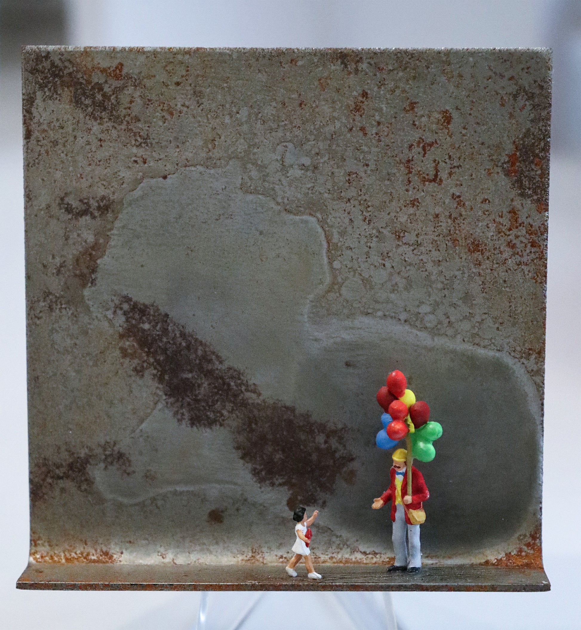 Original Objekt - Assemblage von Katrin Schramm, Titel „Lass sie fliegen...“ – Metallwinkel mit Miniatur-Figuren: Ein Luftballonverkäufer mit vielen bunten Ballons, stehend, hält die  Hand auf und schaut zu einem kleinen Mädchen im weißen Kleid, dass auf  ihn zuläuft. Dahinter das Metall des Winkels mit Struktur, als Hintergrund. Objektcollage, erhältlich bei Galerie Waldbach - What The Artist Made