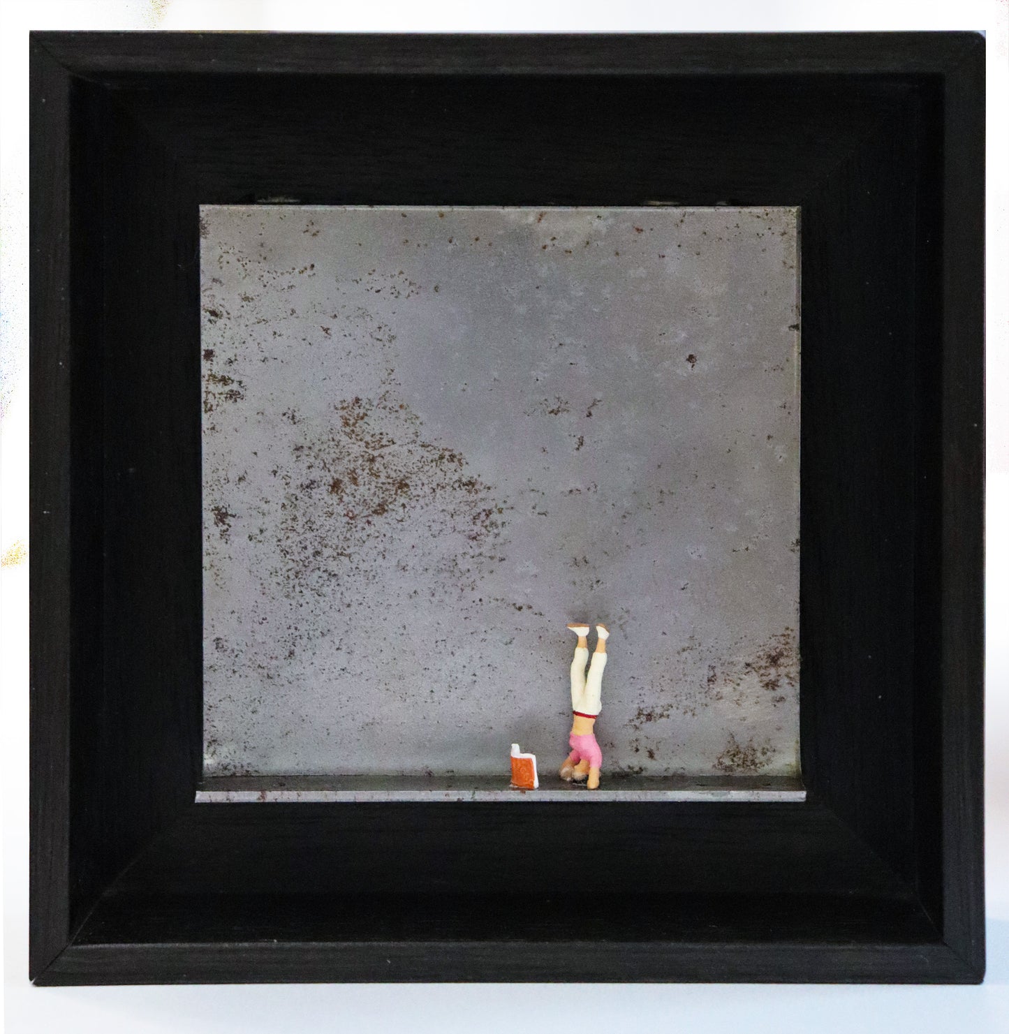 Original Objekt - Assemblage von Katrin Schramm, Titel „180°“ – Metallwinkel mit Miniatur-Figur:  Eine Frau macht einen Kopfstand, vor ihr steht ein Buch aufgeklappt. Dahinter das Metall des Winkels mit Struktur, als Hintergrund. In einem schwarzen Rahmen aus Holz. Objektcollage, erhältlich bei Galerie Waldbach - What The Artist Made