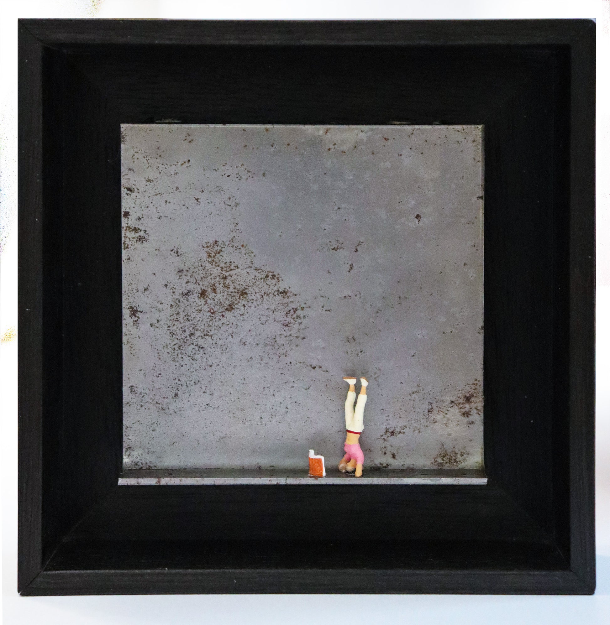 Original Objekt - Assemblage von Katrin Schramm, Titel „180°“ – Metallwinkel mit Miniatur-Figur:  Eine Frau macht einen Kopfstand, vor ihr steht ein Buch aufgeklappt. Dahinter das Metall des Winkels mit Struktur, als Hintergrund. In einem schwarzen Rahmen aus Holz. Objektcollage, erhältlich bei Galerie Waldbach - What The Artist Made