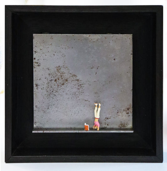 Original Objekt - Assemblage von Katrin Schramm, Titel „180°“ – Metallwinkel mit Miniatur-Figur:  Eine Frau macht einen Kopfstand, vor ihr steht ein Buch aufgeklappt. Dahinter das Metall des Winkels mit Struktur, als Hintergrund. In einem schwarzen Rahmen aus Holz. Objektcollage, erhältlich bei Galerie Waldbach - What The Artist Made