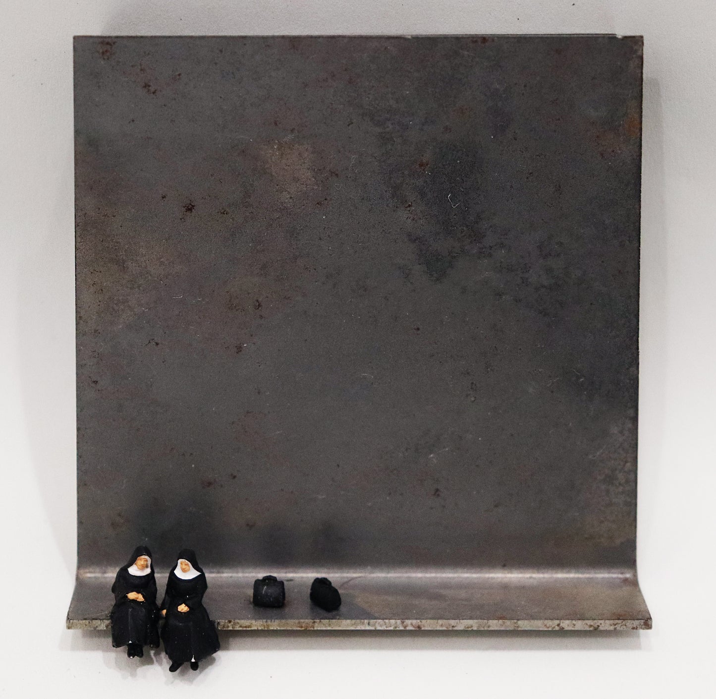 Original Objekt - Assemblage von Katrin Schramm, Titel „Ach Gott...“ – Metallwinkel mit Miniatur-Figuren:  Zwei Nonnen sitzen im schwarz-weißen Habit am linken Rand des Metallwinkels. Rechts von ihnen stehen zwei  schwarze Reisetaschen. Dahinter das Metall des Winkels mit Struktur, als Hintergrund. - Objektcollage, erhältlich bei Galerie Waldbach - What The Artist Made