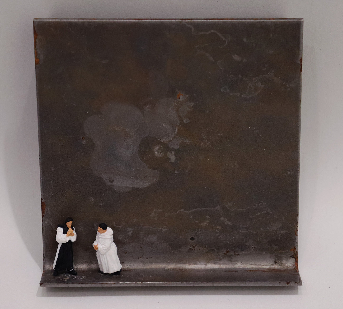 Original Objekt - Assemblage von Katrin Schramm, Titel „Also schweigen wir!“ – Metallwinkel mit Miniatur-Figuren: zwei Priester, einer in schwarzem Habit, einer in weißem Habit, beide stehend, Hände gefaltet. Dahinter das Metall des Winkels mit Struktur, als Hintergrund. - Objektcollage, erhältlich bei Galerie Waldbach - What The Artist Made
