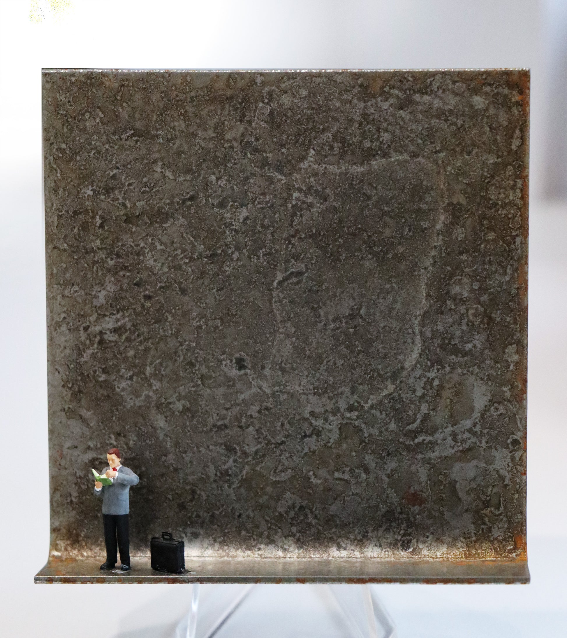 Original Objekt - Assemblage von Katrin Schramm, Titel „Bücher sind Lebensmittel“ – Metallwinkel mit Miniatur-Figur: Ein Mann steht am linken Rand des Winkels und blättert  stehend durch ein Buch, das er hält. Rechts neben ihm steht eine schwarze Aktentasche. Dahinter das Metall des Winkels mit Struktur, als Hintergrund. Objektcollage, erhältlich bei Galerie Waldbach - What The Artist Made