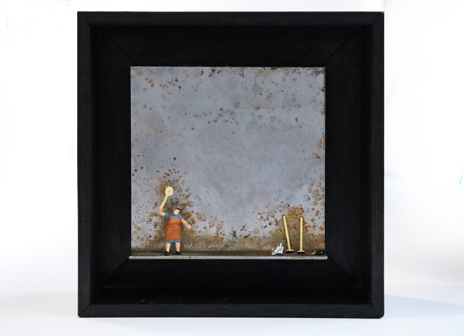 Original Objekt - Assemblage von Katrin Schramm, Titel „Das bisschen Haushalt...“ – Metallwinkel mit Miniatur-Figur: Eine Frau steht in Hausarbeitskleidung am linken Rand des Winkels und hält einen Teppichklopfer hoch. Am rechten Rand stehen zwei Kehrbesen angelehnt, sowie Kehrblech. Dahinter das Metall des Winkels mit Struktur, als Hintergrund. In einem schwarzen Rahmen aus Holz. Objektcollage, erhältlich bei Galerie Waldbach - What The Artist Made