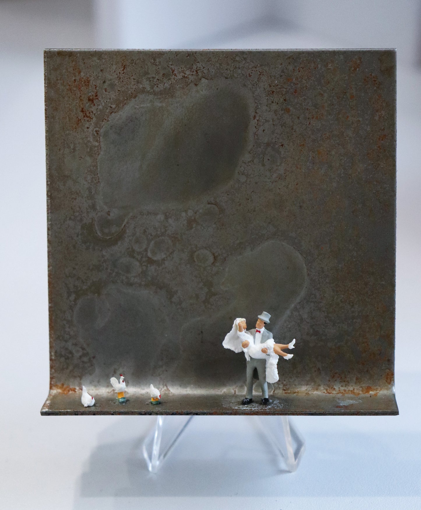Original Objekt - Assemblage von Katrin Schramm, Titel „Dem Himmel so nah...“ – Metallwinkel mit Miniatur-Figuren: In etwa  der Mitte des Winkels steht ein Bräutigam und trägt die Braut in den Armen. Links neben ihnen am Rand stehen drei weiße Hühner. Dahinter das Metall des Winkels mit Struktur, als Hintergrund. Objektcollage, erhältlich bei Galerie Waldbach - What The Artist Made