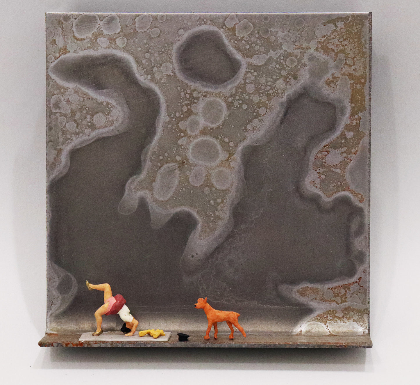 Original Objekt - Assemblage von Katrin Schramm, Titel „Die Lust am Können...“ – Metallwinkel mit Miniatur-Figuren: am linken Bildrand Frau in yoga-ähnlicher Pose/ Brücke machend mit gehobenem Bein, neben ihr ein Zylinder liegend und ein Hund, auf sie zugehend. Dahinter das Metall des Winkels mit Struktur, als Hintergrund. - Objektcollage, erhältlich bei Galerie Waldbach - What The Artist Made