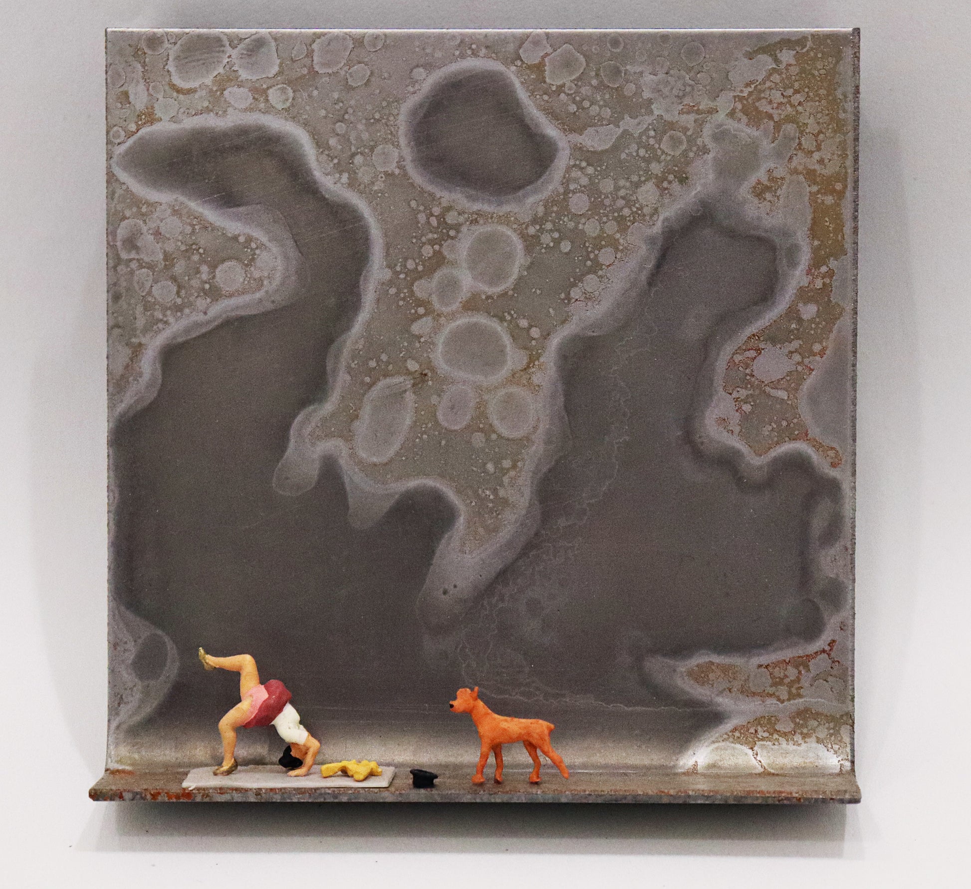Original Objekt - Assemblage von Katrin Schramm, Titel „Die Lust am Können...“ – Metallwinkel mit Miniatur-Figuren: am linken Bildrand Frau in yoga-ähnlicher Pose/ Brücke machend mit gehobenem Bein, neben ihr ein Zylinder liegend und ein Hund, auf sie zugehend. Dahinter das Metall des Winkels mit Struktur, als Hintergrund. - Objektcollage, erhältlich bei Galerie Waldbach - What The Artist Made