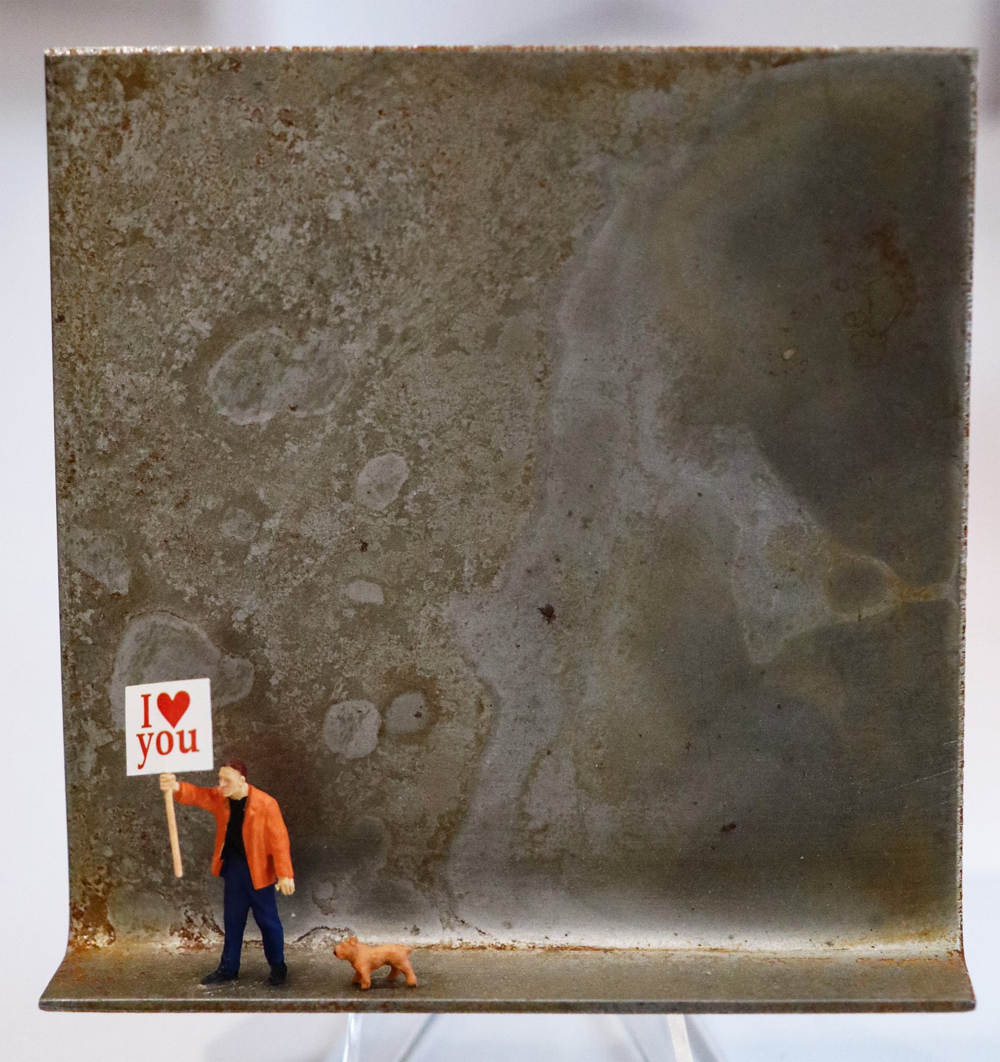 Original Objekt - Assemblage von Katrin Schramm, Titel „Einfach so...“ – Metallwinkel mit Miniatur-Figuren:  Eine Person steht am linken Bildrand in einer orangenen Jacke mit einem Schild. neben ihr ein kleiner Hund. Auf dem Schild  steht "I love you", das love als Herz dargestellt. Dahinter das Metall des Winkels mit Struktur, als Hintergrund. - Objektcollage, erhältlich bei Galerie Waldbach - What The Artist Made