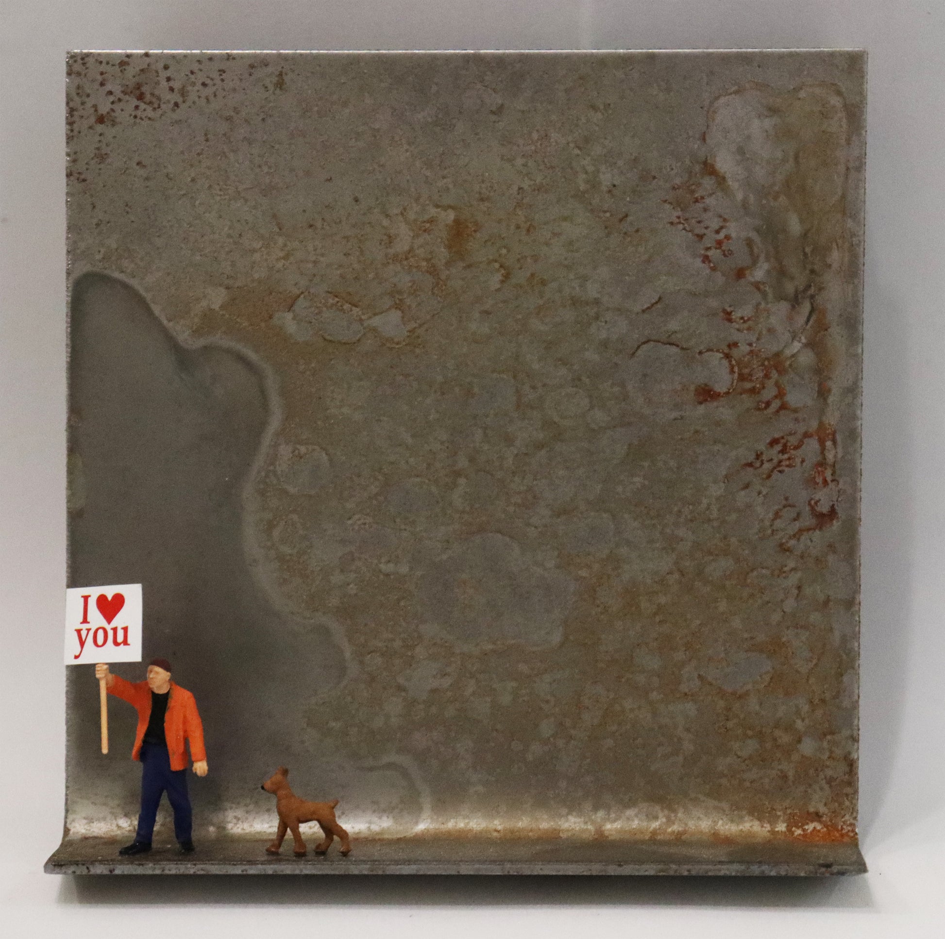 Original Objekt - Assemblage von Katrin Schramm, Titel „Einfach so“ – Metallwinkel mit Miniatur-Figuren: Eine Person (mann) in orangener Jacke am linken Bildrand stehend, daneben ein Hund. Der Mann hält ein Schild hoch mit der Beschriftung "I love (heart) you. Dahinter das Metall des Winkels mit Struktur, als Hintergrund. - Objektcollage, erhältlich bei Galerie Waldbach - What The Artist Made
