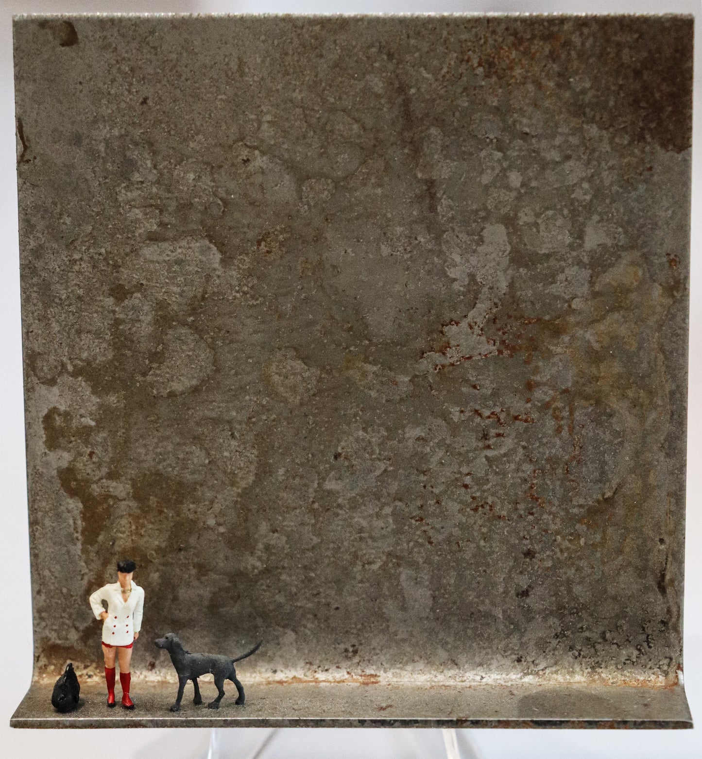 Original Objekt - Assemblage von Katrin Schramm, Titel „Geduld ist der Seele Schild...“ – Metallwinkel mit Miniatur-Figuren: Eine Frau in weißem Jacket mit roten Stiefeln am linken Rand stehend. Rechts neben ihr ist ein großer dunkelgrauer Hund, links von ihr eine große schwarze Tragetasche. Dahinter das Metall des Winkels mit Struktur, als Hintergrund. - Objektcollage, erhältlich bei Galerie Waldbach - What The Artist Made