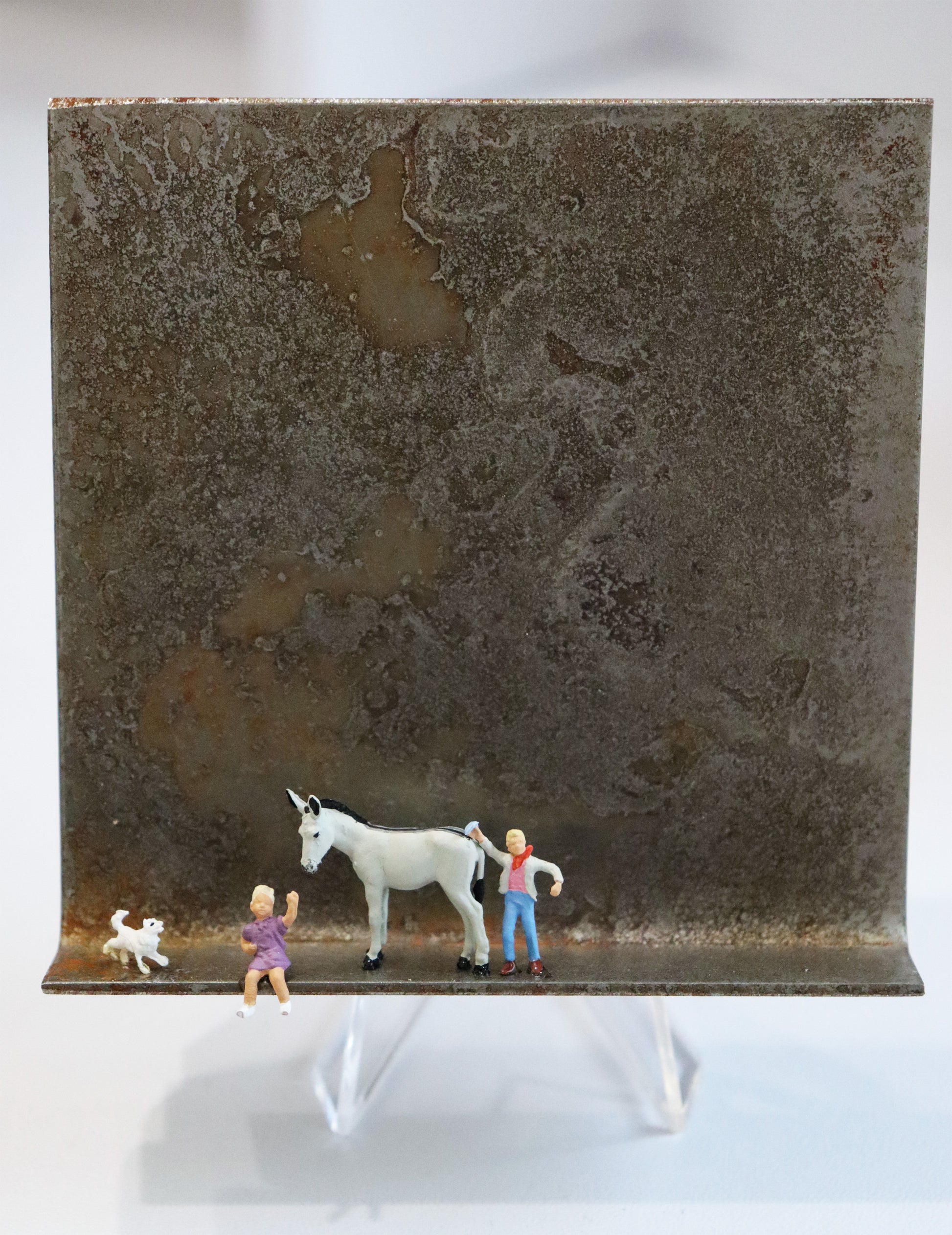 Original Objekt - Assemblage von Katrin Schramm, Titel „Mit Eselsgeduld...“ – Metallwinkel mit Miniatur-Figuren: Ein weißer Esel stehend, rechts von ihm steht ein Junge und hebt die Hand, vor dem Esel sitzt ein Mädchen und winkt. Neben beiden am linken Rand des Winkels ist ein kleiner weißer Hund. Dahinter das Metall des Winkels mit Struktur, als Hintergrund. Objektcollage, erhältlich bei Galerie Waldbach - What The Artist Made