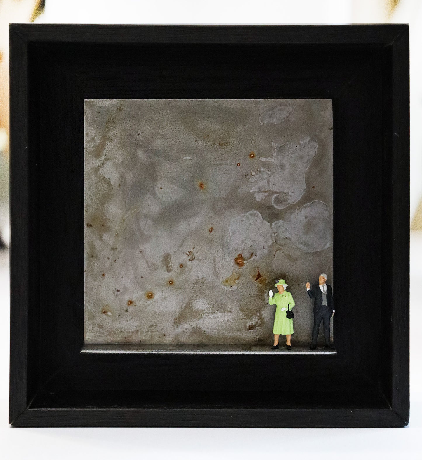 Original Objekt - Assemblage von Katrin Schramm, Titel „Never dressed down...“ – Metallwinkel mit Miniatur-Figuren:  Am rechten Rand stehen Queen Elisabeth II und Prinz Philip, beide winken. Queen Elisabeth trägt ein hellgrünes Kostüm. Dahinter das Metall des Winkels mit Struktur, als Hintergrund. In einem schwarzen Rahmen aus Holz. Objektcollage, erhältlich bei Galerie Waldbach - What The Artist Made