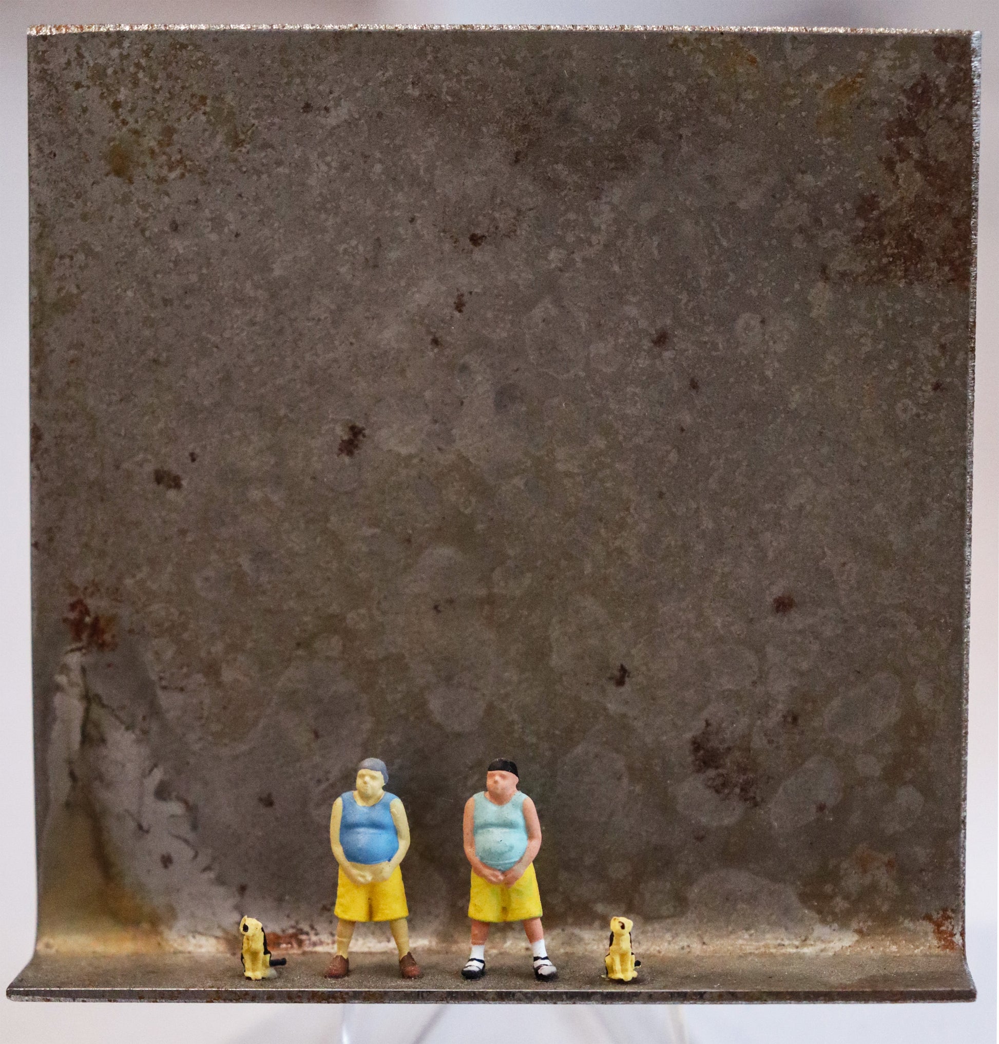 Original Objekt - Assemblage von Katrin Schramm, Titel „Teambuilding“ – Metallwinkel mit Miniatur-Figuren: Zwei Männer stehen breitbeinig mittig da und halten sich das Genital, wie zum Wasserlassen. Links und Rechts von ihnen sitzt jeweils ein beigefarbener Hund. Dahinter das Metall des Winkels mit Struktur, als Hintergrund. - Objektcollage, erhältlich bei Galerie Waldbach - What The Artist Made
