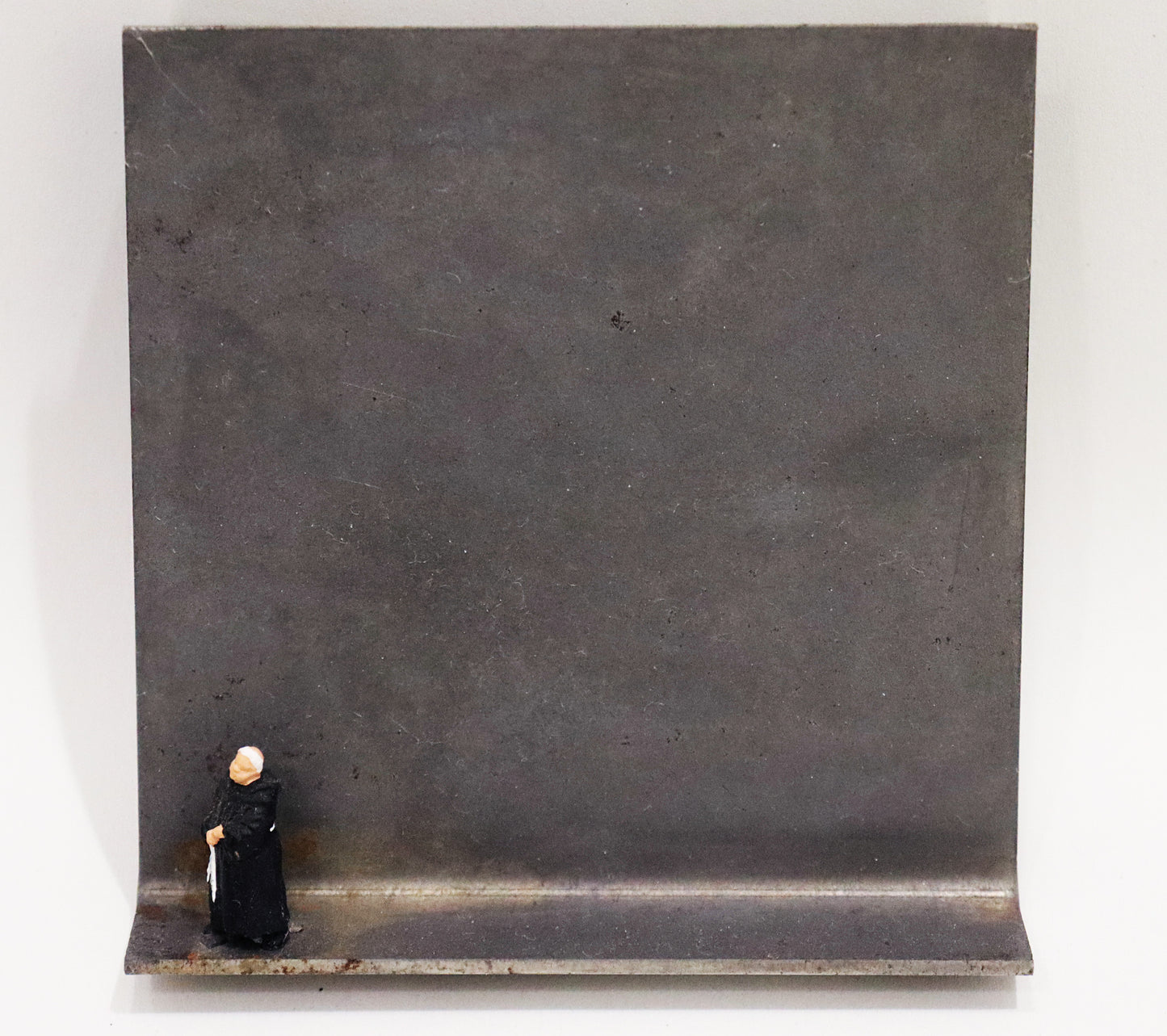 Original Objekt - Assemblage von Katrin Schramm, Titel „Wenn ich stehe, dann stehe ich.“ – Metallwinkel mit Miniatur-Figur: am linken Bildrand Mönch in schwarzem Habit stehend, mit gefalteten Händen und zur Seite schauend. Dahinter das Metall des Winkels mit Struktur, als Hintergrund. - Objektcollage, erhältlich bei Galerie Waldbach - What The Artist Made