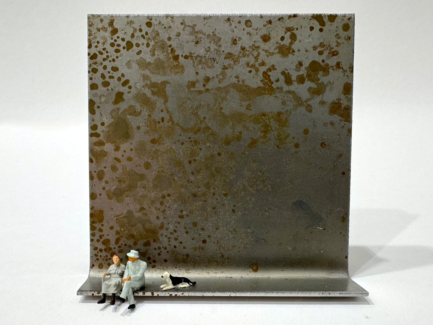 Original Objekt - Assemblage von Katrin Schramm, Titel „Wolke 9“ – Metallwinkel mit Miniatur-Figuren: zwei Personen am linken Rand des Metallwinkels sitzend, davon eine mit Gehstock. Daneben ein schwarz weißer Hund, dahinter das Metall des Winkels als Hintergrund. - Objektcollage, erhältlich bei What The Artist Made