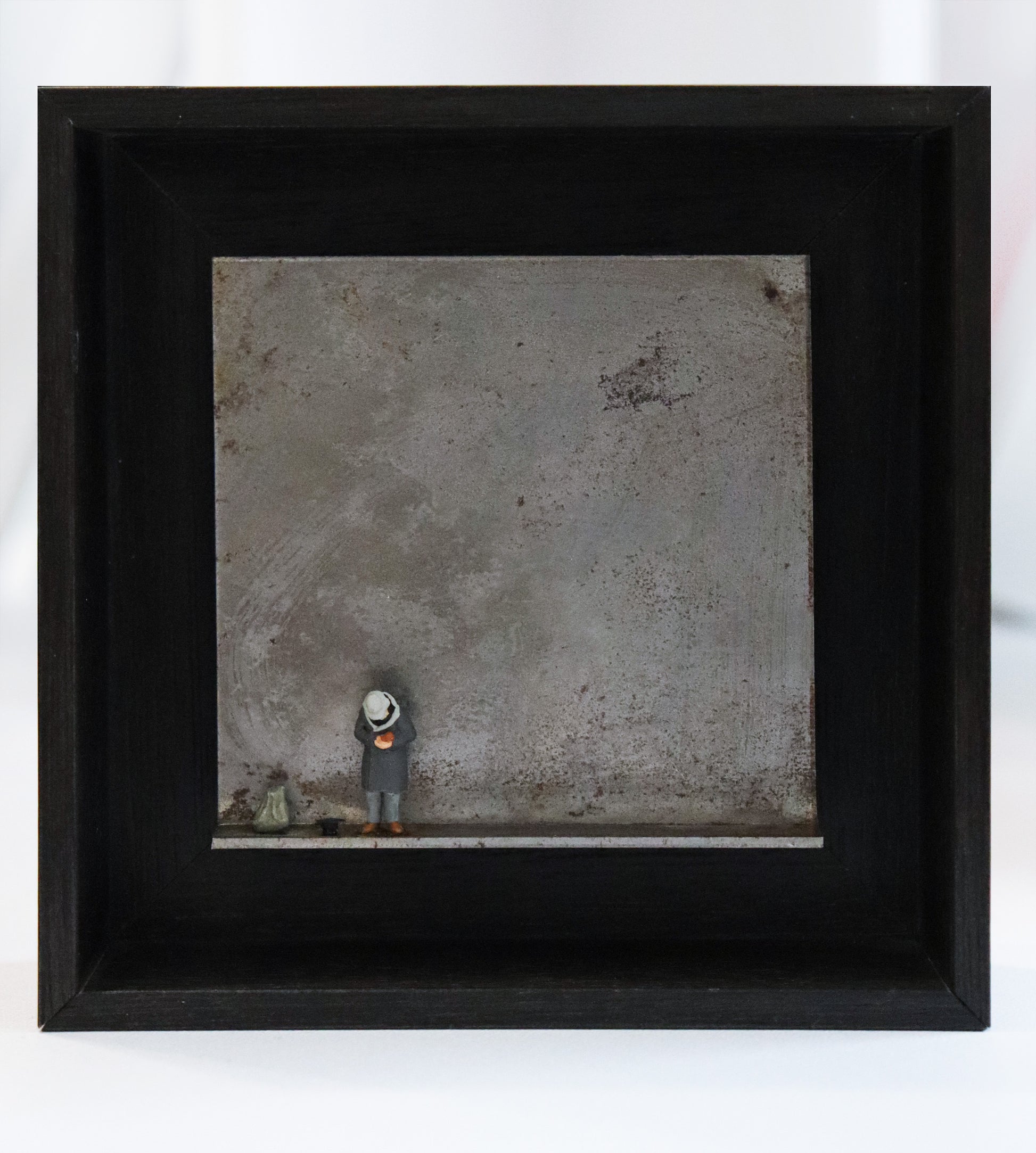 Original Objekt - Assemblage von Katrin Schramm, Titel „geben...“ – Metallwinkel mit Miniatur-Figur: Ein Mann im grauen Mantel steht  am linken Rand des Winkels, der Kopf gebeugt, unter einem Hut nicht sichtbar. Neben ihm steht ein schwarzer Hut auf dem Boden und ein Beutel. Dahinter das Metall des Winkels mit Struktur, als Hintergrund. In einem schwarzen Rahmen aus Holz. Objektcollage, erhältlich bei Galerie Waldbach - What The Artist Made