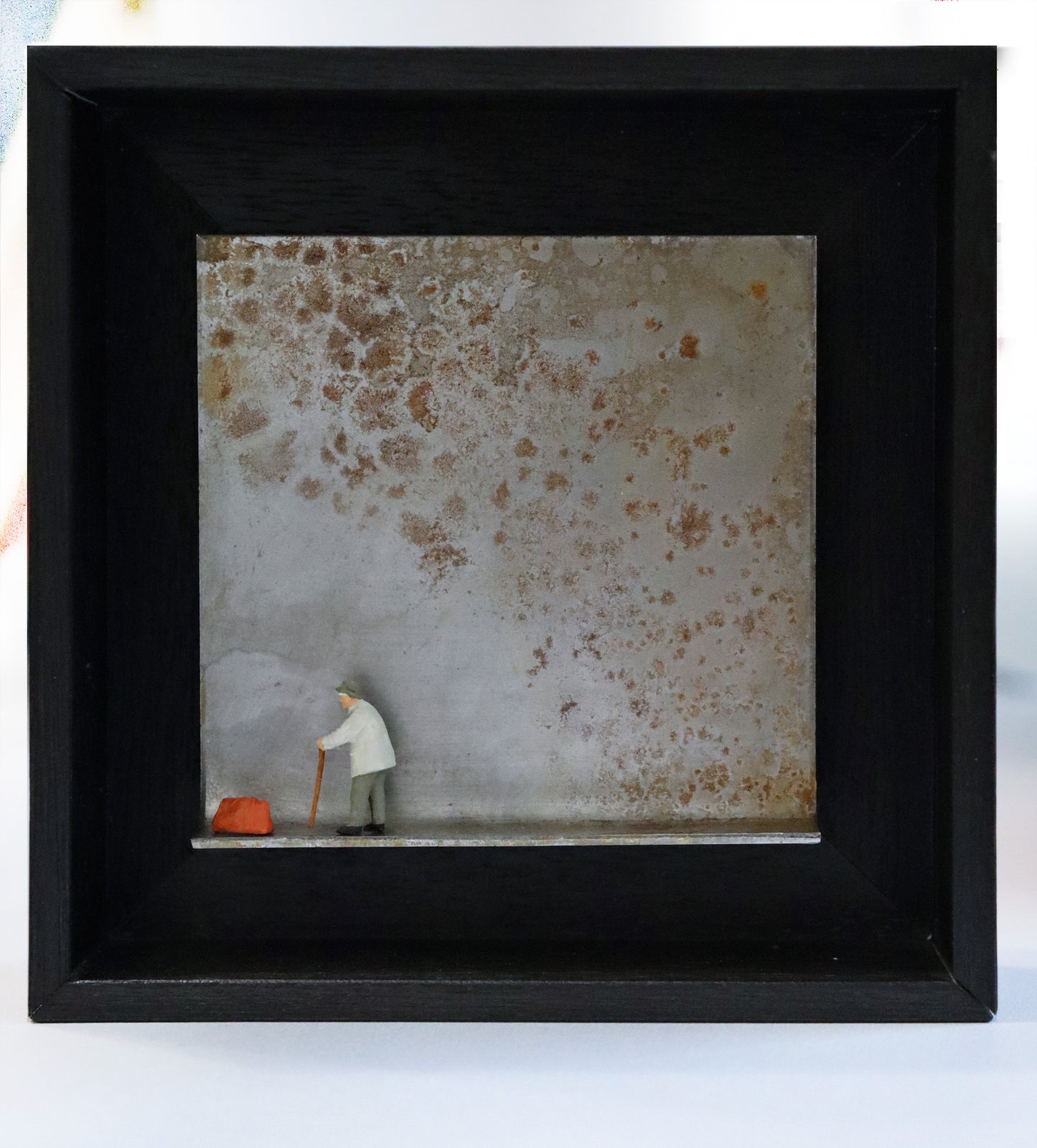 Original Objekt - Assemblage von Katrin Schramm, Titel „gebeugt, nicht gebrochen“ – Metallwinkel mit Miniatur-Figur: Am linken Rand steht eine ältere Person, auf einem Gehstock gebeugt, in grauer Kleidung. Vor ihr eine orange Reisetasche stehend. Dahinter das Metall des Winkels mit Struktur, als Hintergrund. In einem schwarzen Rahmen aus Holz. Objektcollage, erhältlich bei Galerie Waldbach - What The Artist Made