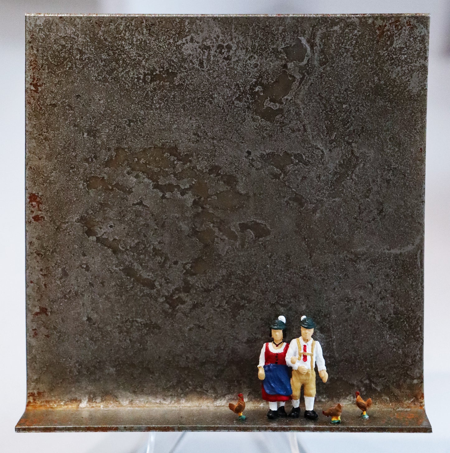 Original Objekt - Assemblage von Katrin Schramm, Titel „Mia san mia“ – Metallwinkel mit Miniatur-Figuren: Zwei Personen (Frau und Mann) stehen rechts der Winkelmitte, um sie herum drei Hühner. Sie tragen Trachtenkleidung und entsprechende Hüte. Dahinter das Metall des Winkels mit Struktur, als Hintergrund. - Objektcollage, erhältlich bei Galerie Waldbach - What The Artist Made
