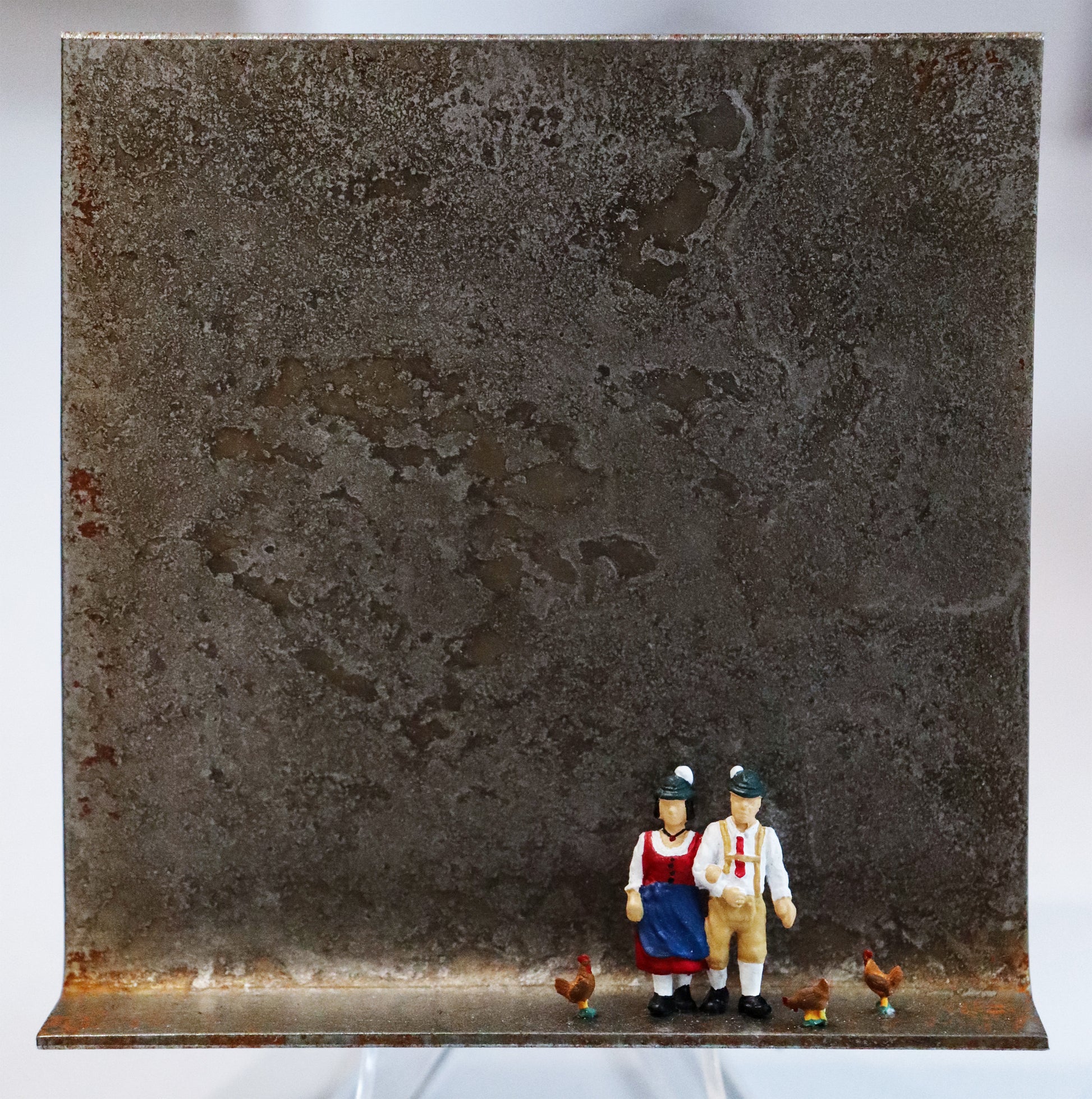 Original Objekt - Assemblage von Katrin Schramm, Titel „Mia san mia“ – Metallwinkel mit Miniatur-Figuren: Zwei Personen (Frau und Mann) stehen rechts der Winkelmitte, um sie herum drei Hühner. Sie tragen Trachtenkleidung und entsprechende Hüte. Dahinter das Metall des Winkels mit Struktur, als Hintergrund. - Objektcollage, erhältlich bei Galerie Waldbach - What The Artist Made