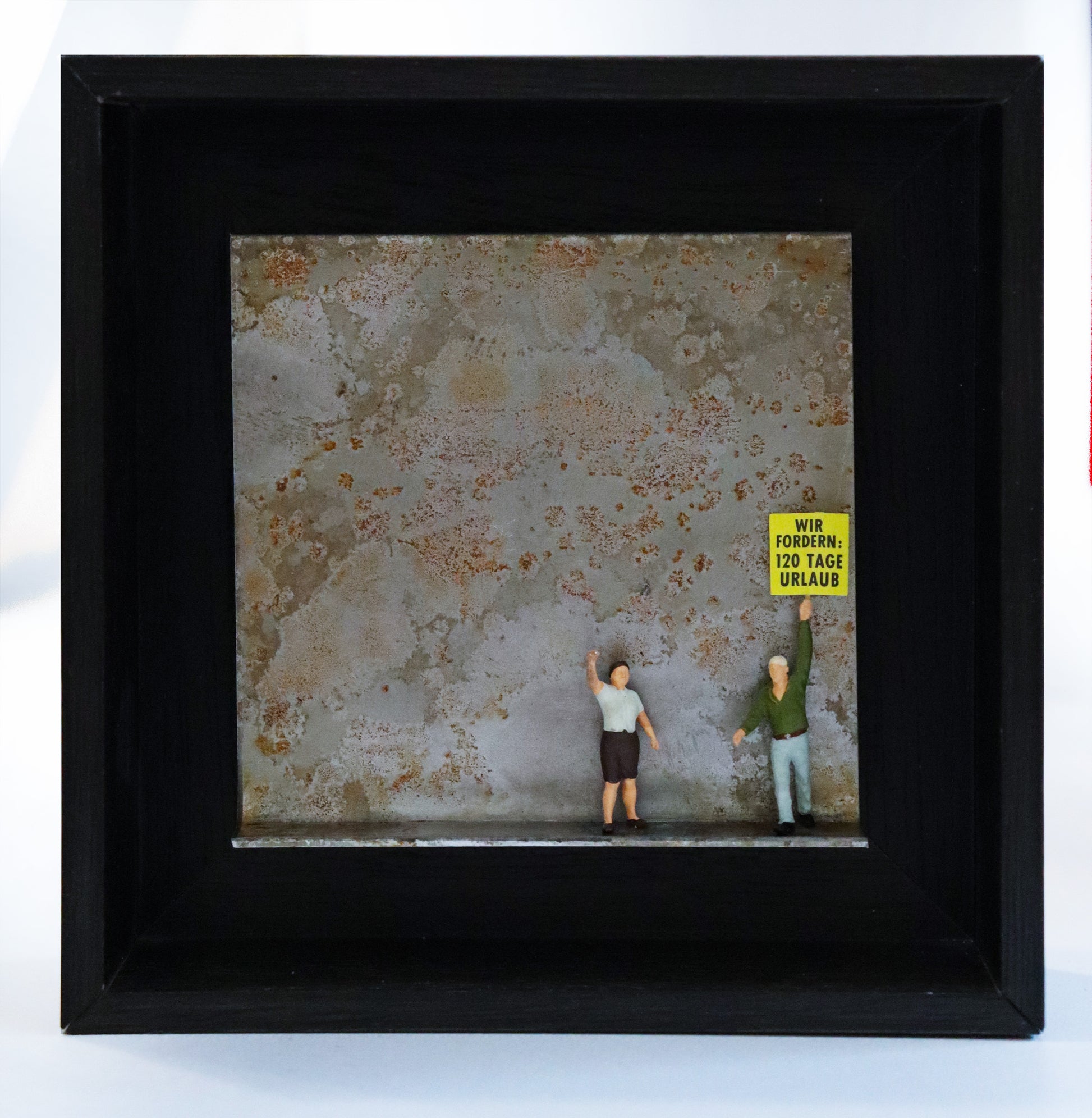Original Objekt - Assemblage von Katrin Schramm, Titel „sind dann mal weg...“ – Metallwinkel mit Miniatur-Figuren: Zwei Personen stehen rechts auf dem Winkel. Die linke Person streckt einen Arm mit Faust hoch, die andere trägt ein gelbes Schild - Anmutung einer Demonstration. Auf dem Schild steht: "Wir fordern: 120 Tage Urlaub". Dahinter das Metall des Winkels mit Struktur, als Hintergrund. In einem schwarzen Rahmen aus Holz. Objektcollage, erhältlich bei Galerie Waldbach - What The Artist Made
