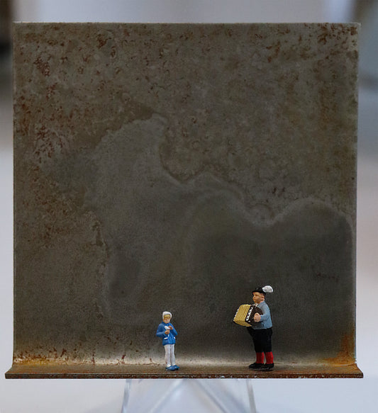 Original Objekt - Assemblage von Katrin Schramm, Titel „Nur a Quetschn ?“ – Metallwinkel mit Miniatur-Figuren: Ein Mann in Tracht mit Akkordeon steht auf der rechten Seite des Winkels. In der Mitte steht ein Kind, etwas in der Hand haltend. Dahinter das Metall des Winkels mit Struktur, als Hintergrund. Objektcollage, erhältlich bei Galerie Waldbach - What The Artist Made