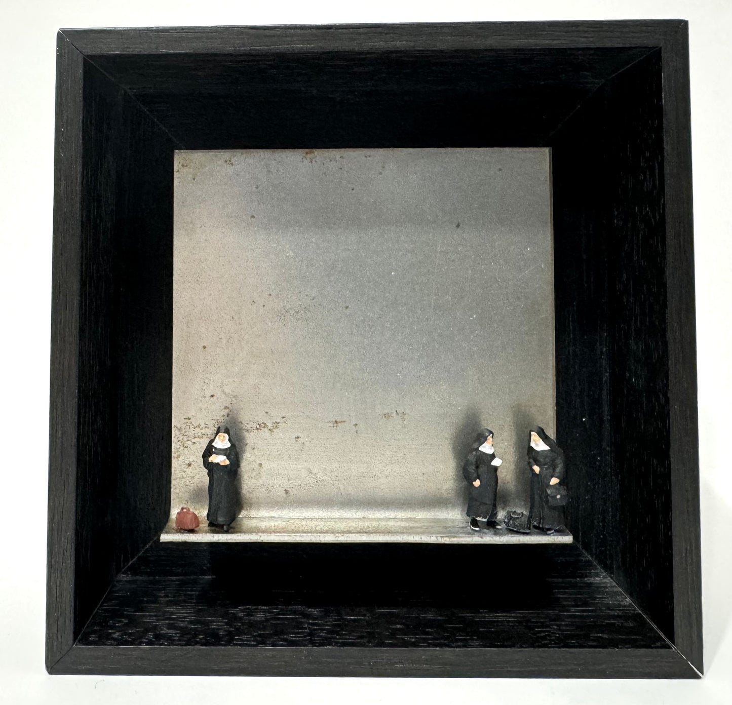 Original Objekt - Assemblage von Katrin Schramm, Titel „auf dem Weg…“ – Metallwinkel mit Miniatur-Figuren: drei Nonnen, davon eine links am Rand des Winkels stehend, zwei rechts am Rand stehend, jeweils mit Reisetaschen. Dahinter das Metall des Winkels als Hintergrund. - Objektcollage, erhältlich bei What The Artist Made