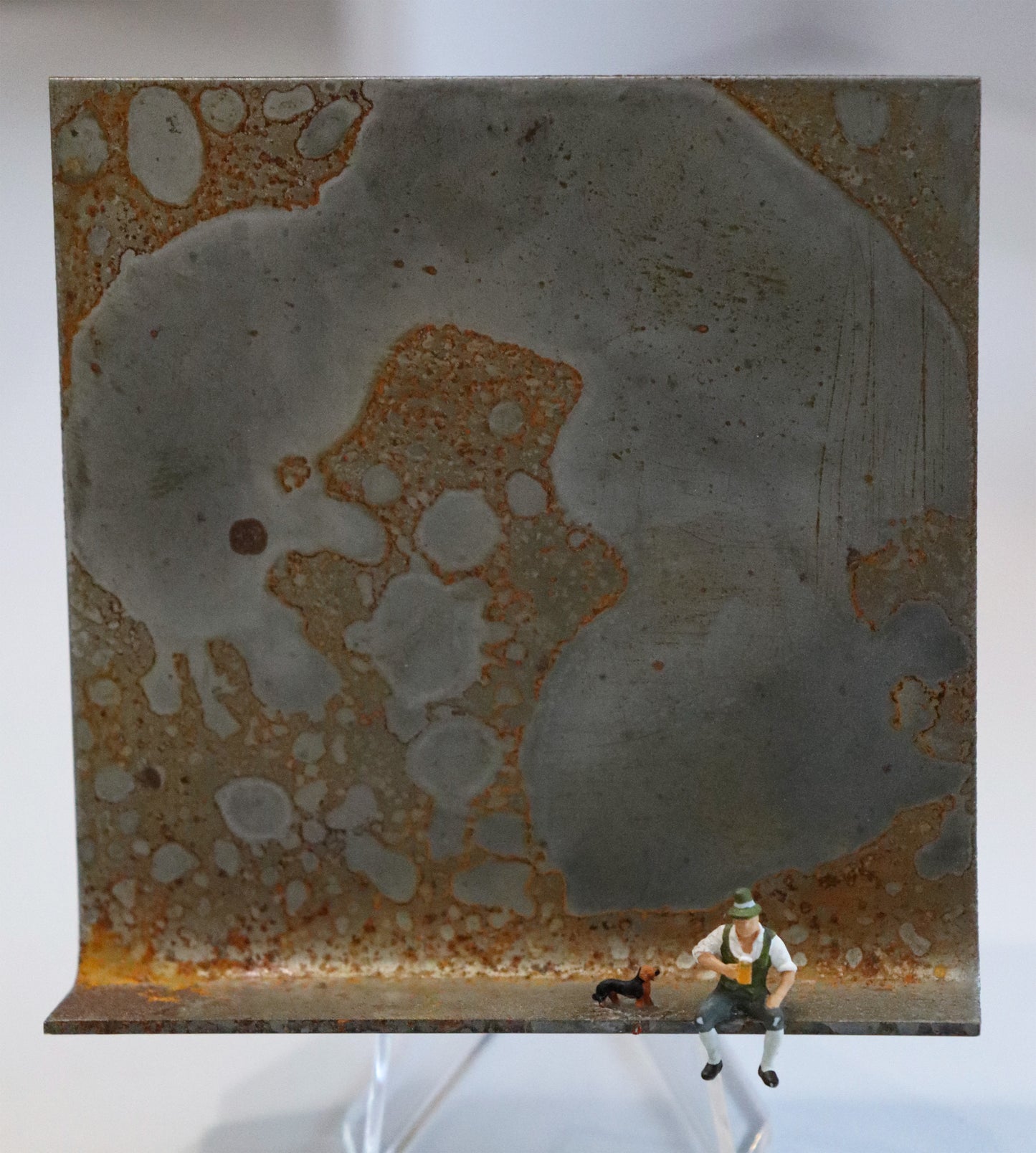 Original Objekt - Assemblage von Katrin Schramm, Titel „Prosit. Zamperl!“ – Metallwinkel mit Miniatur-Figuren: Ein Mann sitzt in Tracht und Lederhose am rechten Winkelrand und hält eine Maß Bier in der Hand. Links neben ihm steht ein Dackelhund. Dahinter das Metall des Winkels mit Struktur, als Hintergrund. Objektcollage, erhältlich bei Galerie Waldbach - What The Artist Made