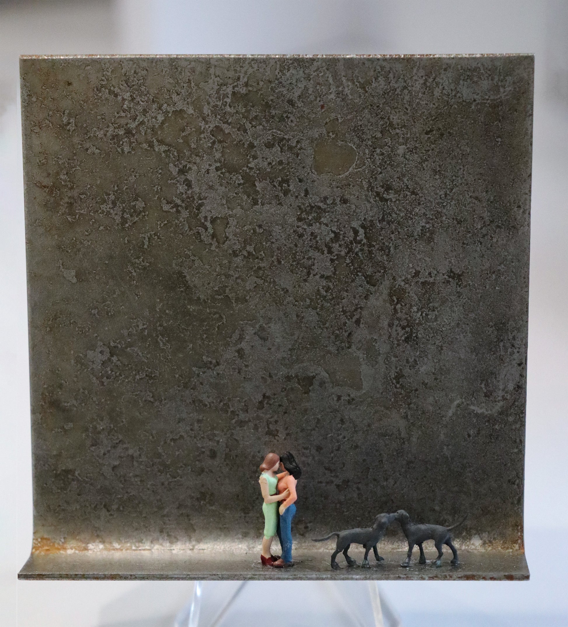 Original Objekt - Assemblage von Katrin Schramm, Titel „bonding“ – Metallwinkel mit Miniatur-Figuren: Zwei Frauen stehen mittig  auf dem Winkel und halten sich fest in den Armen, sie berühren sich an der Stirn. Rechts neben ihnen stehen zwei große graue Hunde und berühren sich an den Schnauzen. Dahinter das Metall des Winkels mit Struktur, als Hintergrund. Objektcollage, erhältlich bei Galerie Waldbach - What The Artist Made